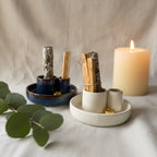 Two ceramic smudge kit holders with white and dark blue glazes, containing bundles of dried herbs and sage sticks, arranged on a textured white fabric surface with a blurred background.