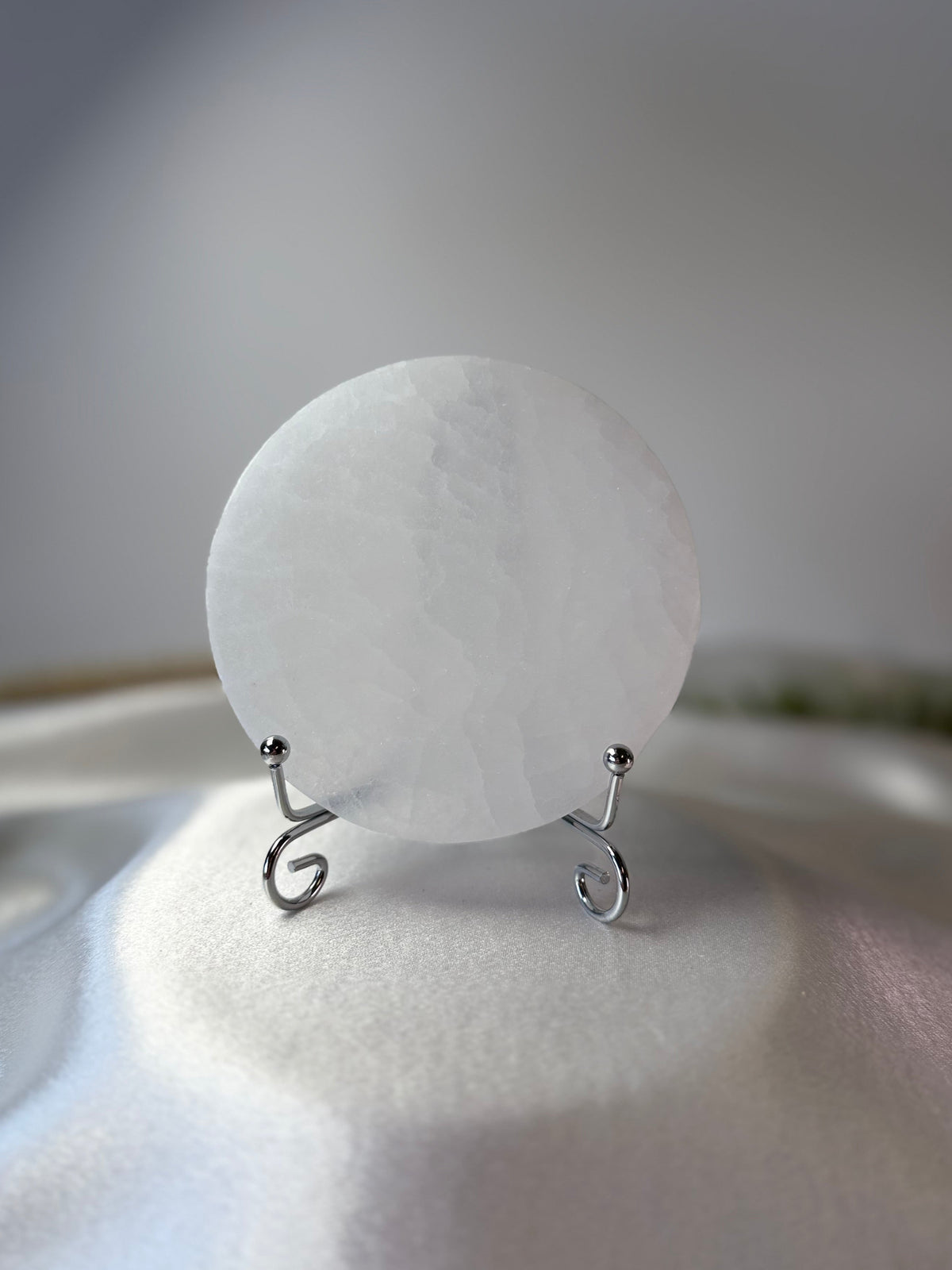 A round, translucent white crystal or gemstone plaque displayed on a small silver wire stand against a soft, light gray background.
