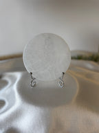 A round, translucent white stone or glass disc displayed on a small silver wire stand, set against a soft-focus background with draped satin fabric.