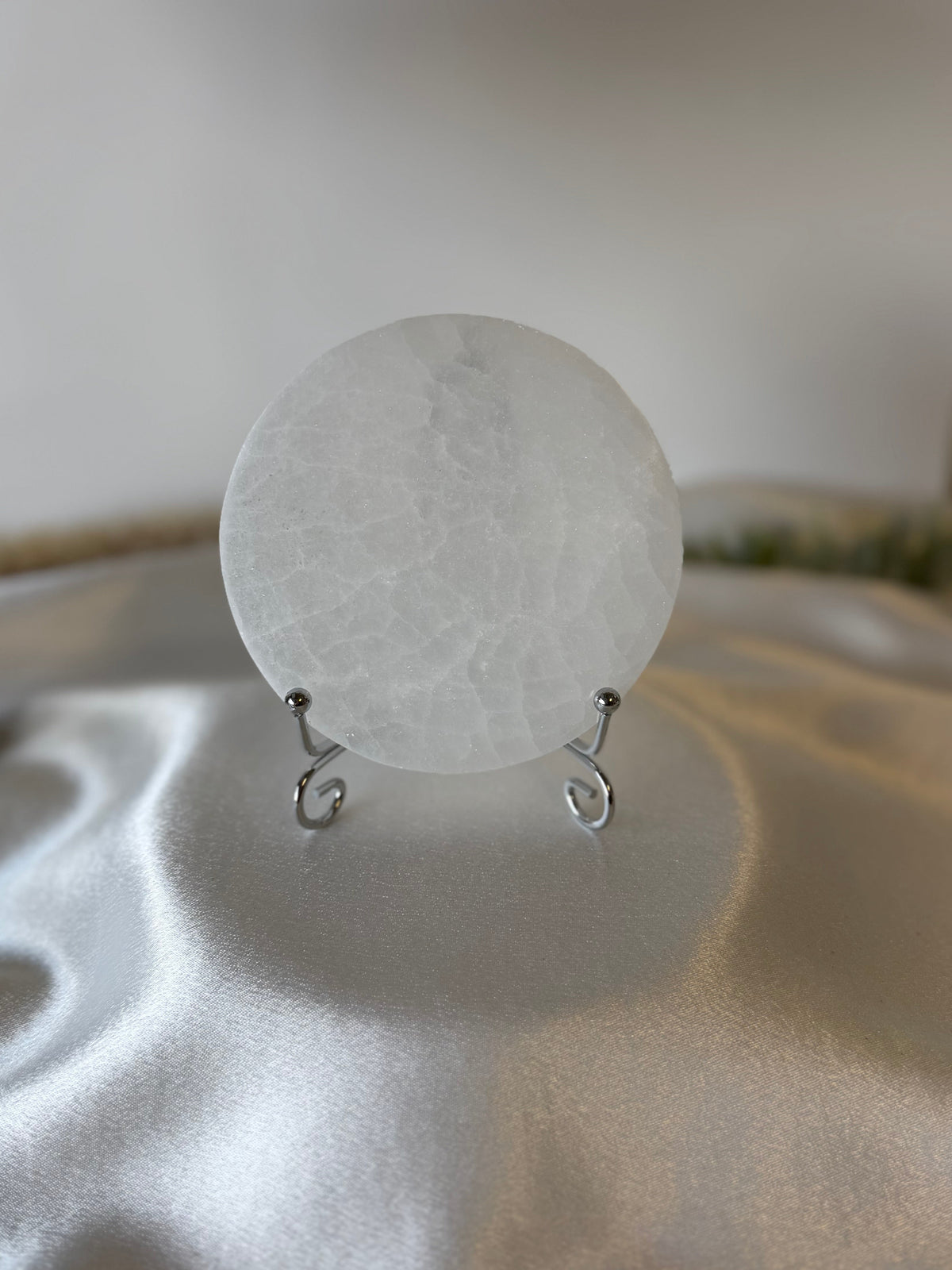 A round, translucent white stone or glass disc displayed on a small silver wire stand, set against a soft-focus background with draped satin fabric.