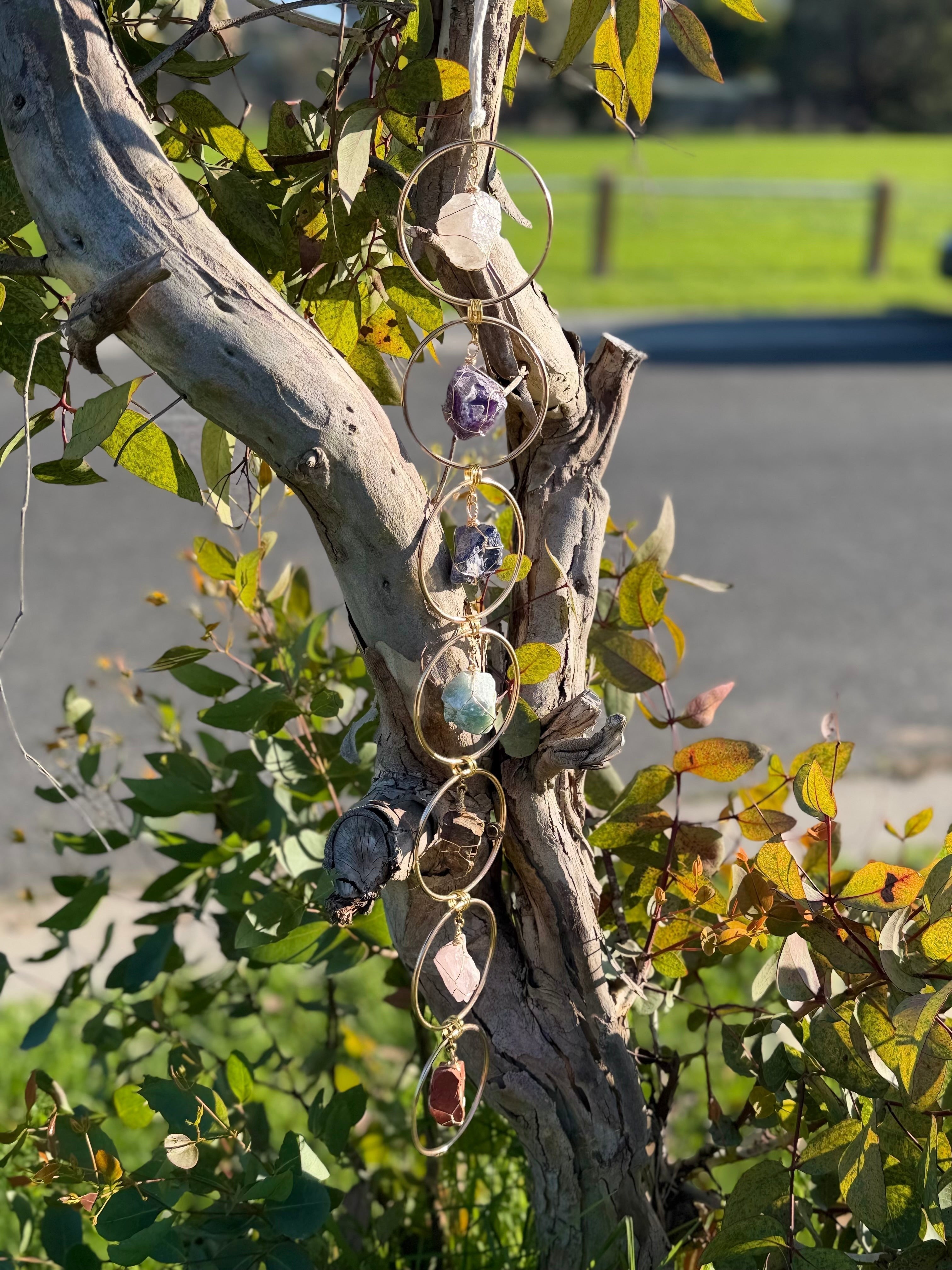 Treasured Energies 7 chakra crystal window hanging featuring raw gemstones in gold circles hanging from a tree branch.