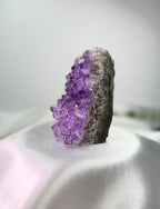 Treasured Energies amethyst cluster with deep purple crystals on a natural stone base, side view on white fabric.
