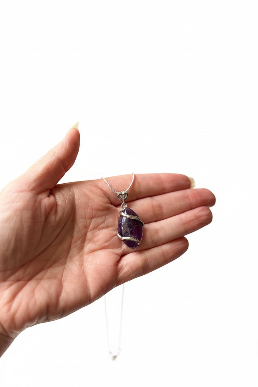 Treasured Energies amethyst natural crystal pendant wrapped in silver wire, held in a palm against a white background.