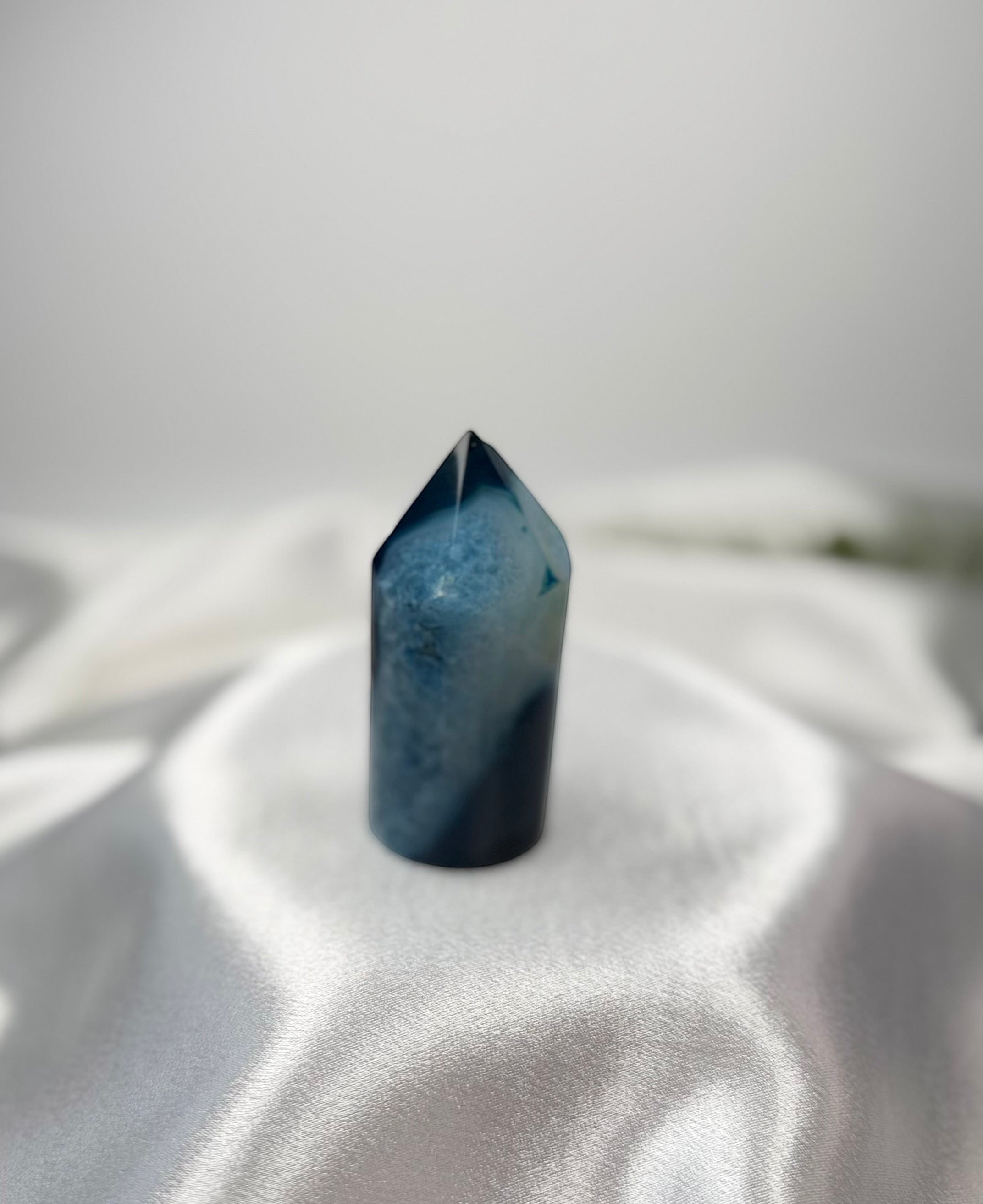 Treasured Energies blue agate crystal tower with a polished point, displayed on a soft white satin fabric.
