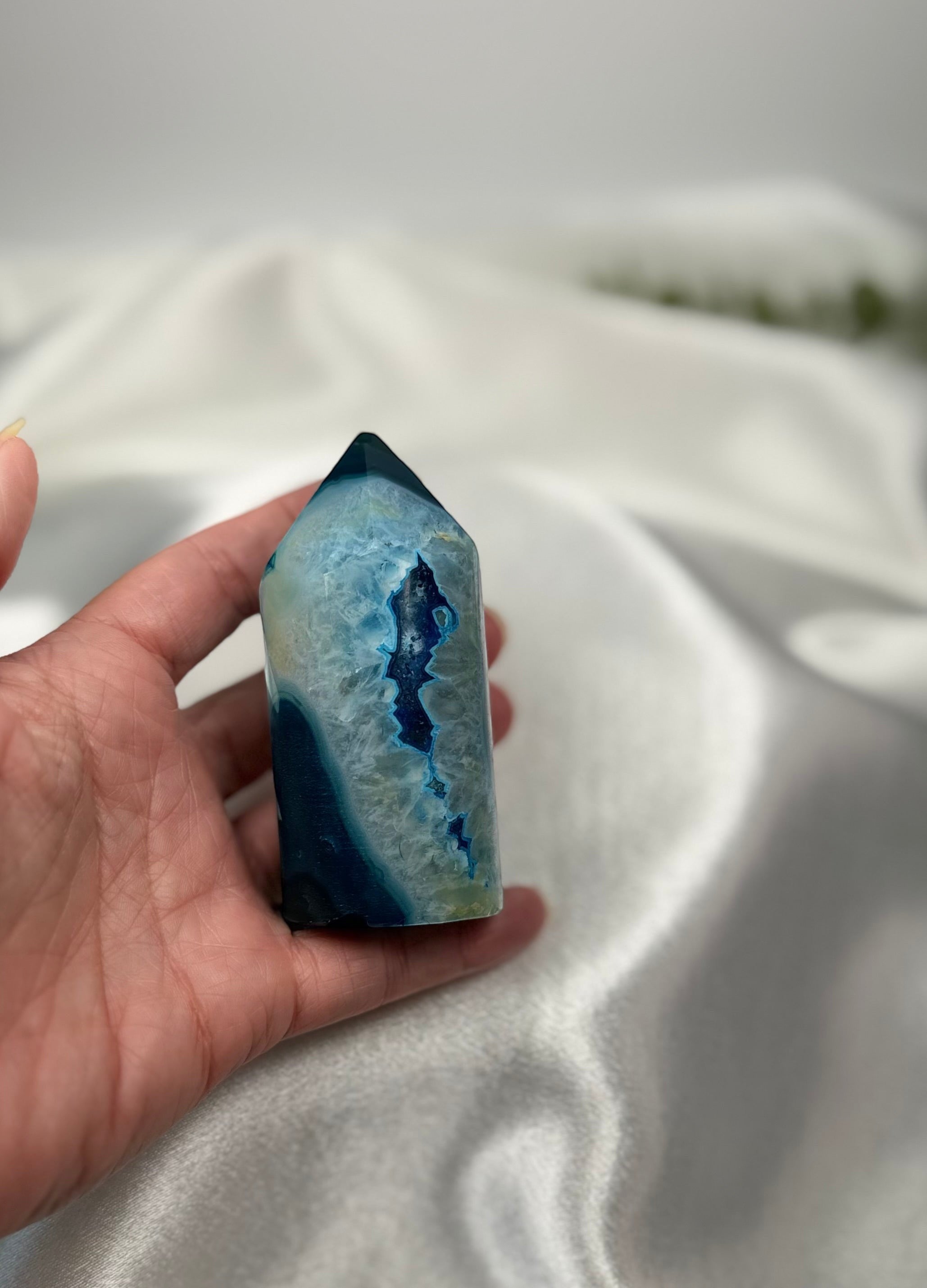 Treasured Energies blue agate crystal tower with a polished point held in a hand against a soft white fabric background
