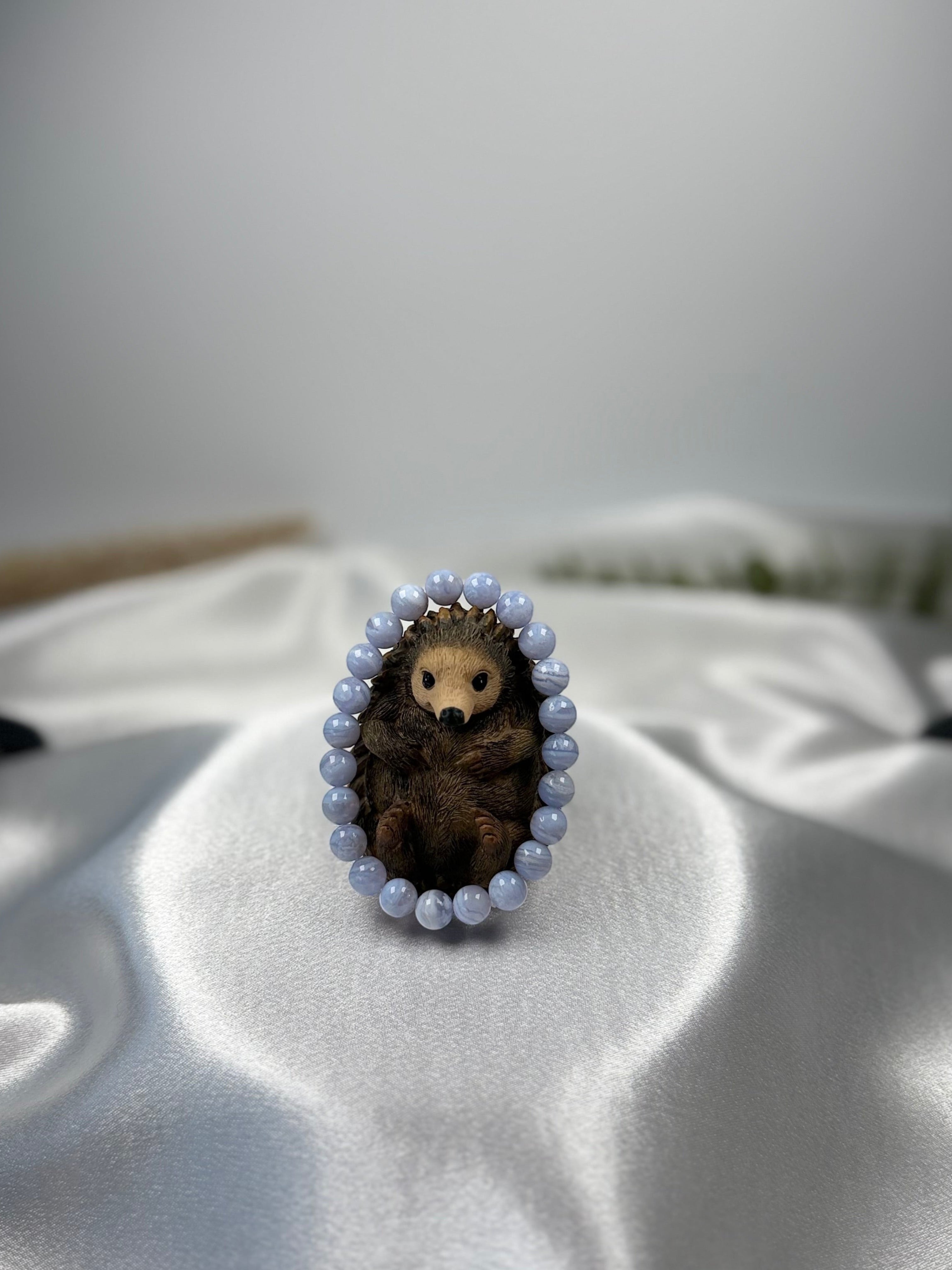 Treasured Energies blue lace agate crystal bead bracelet encircling a small brown hedgehog figurine on satin fabric