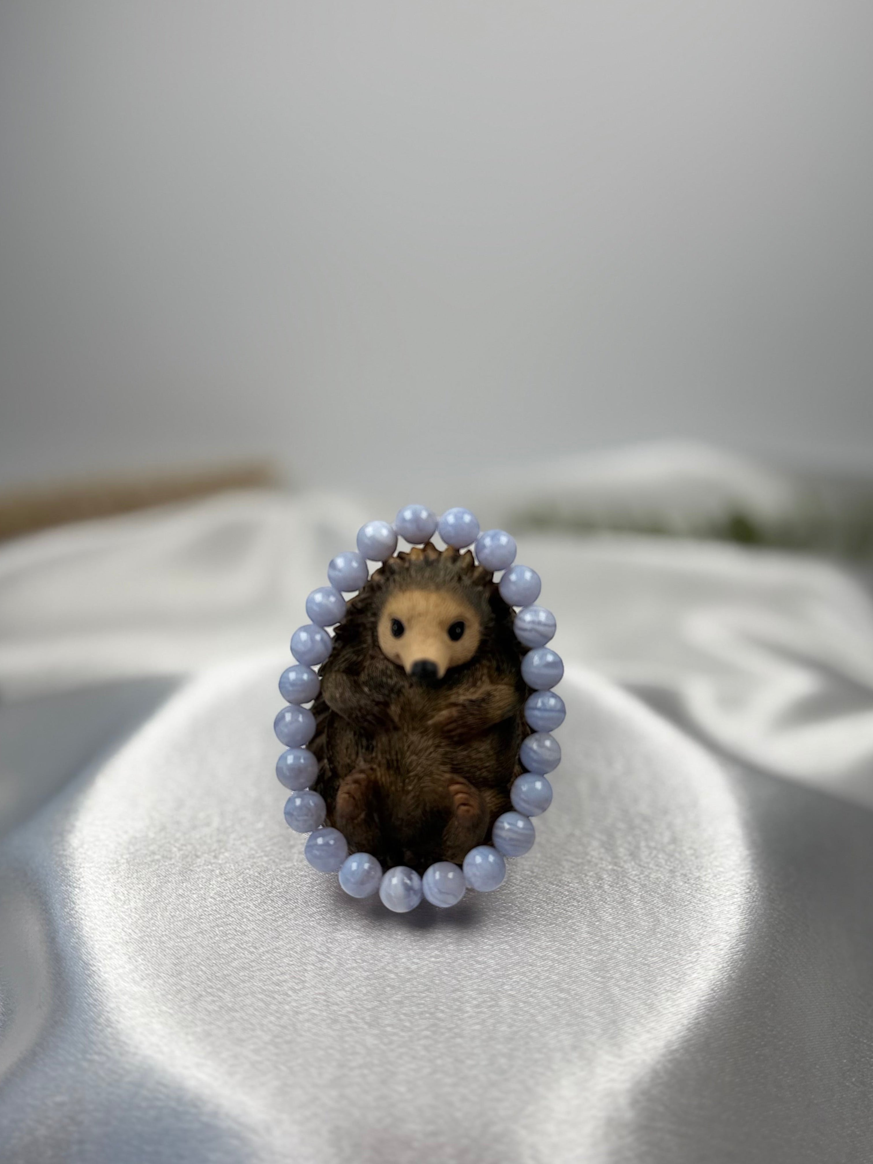 Treasured Energies blue lace agate crystal bead bracelet encircling a small decorative hedgehog figurine on satin
