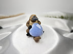 Treasured Energies Blue Lace Agate crystal heart held by a small platypus figurine on a white satin background.