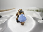 Treasured Energies blue lace agate crystal heart held by a small platypus figurine on a white satin fabric background.
