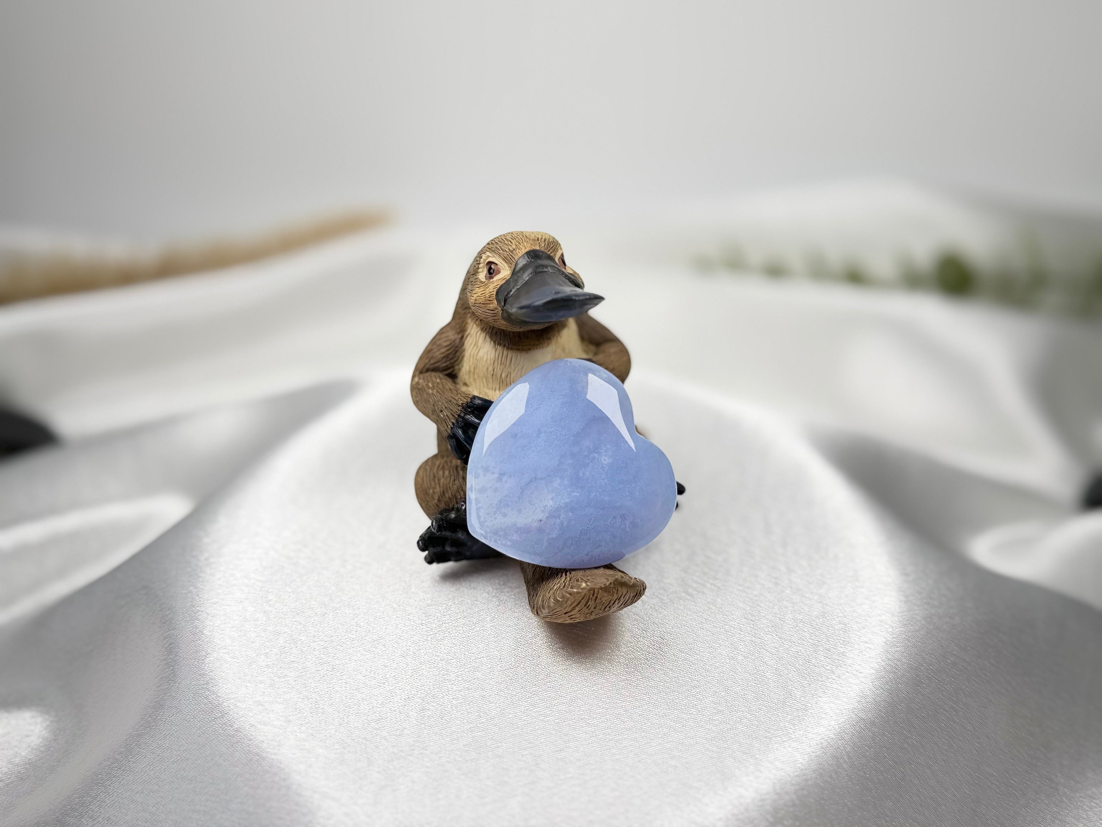Treasured Energies blue lace agate crystal heart held by a small platypus figurine on a white satin fabric background.