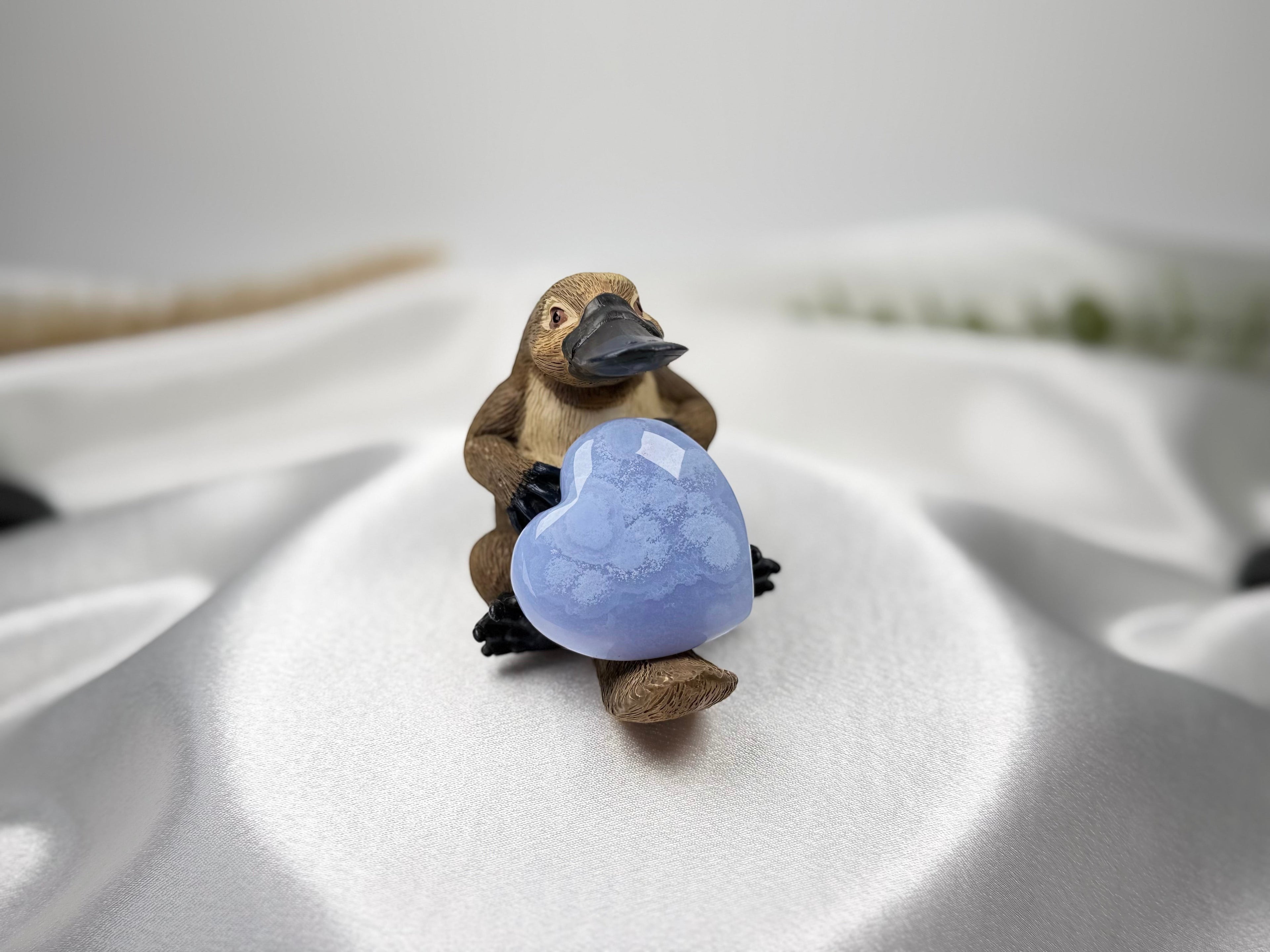 Treasured Energies blue lace agate crystal heart held by a small platypus figurine on a white satin background.