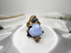 Treasured Energies Blue Lace Agate crystal heart held by a small platypus figurine on a soft white satin background.