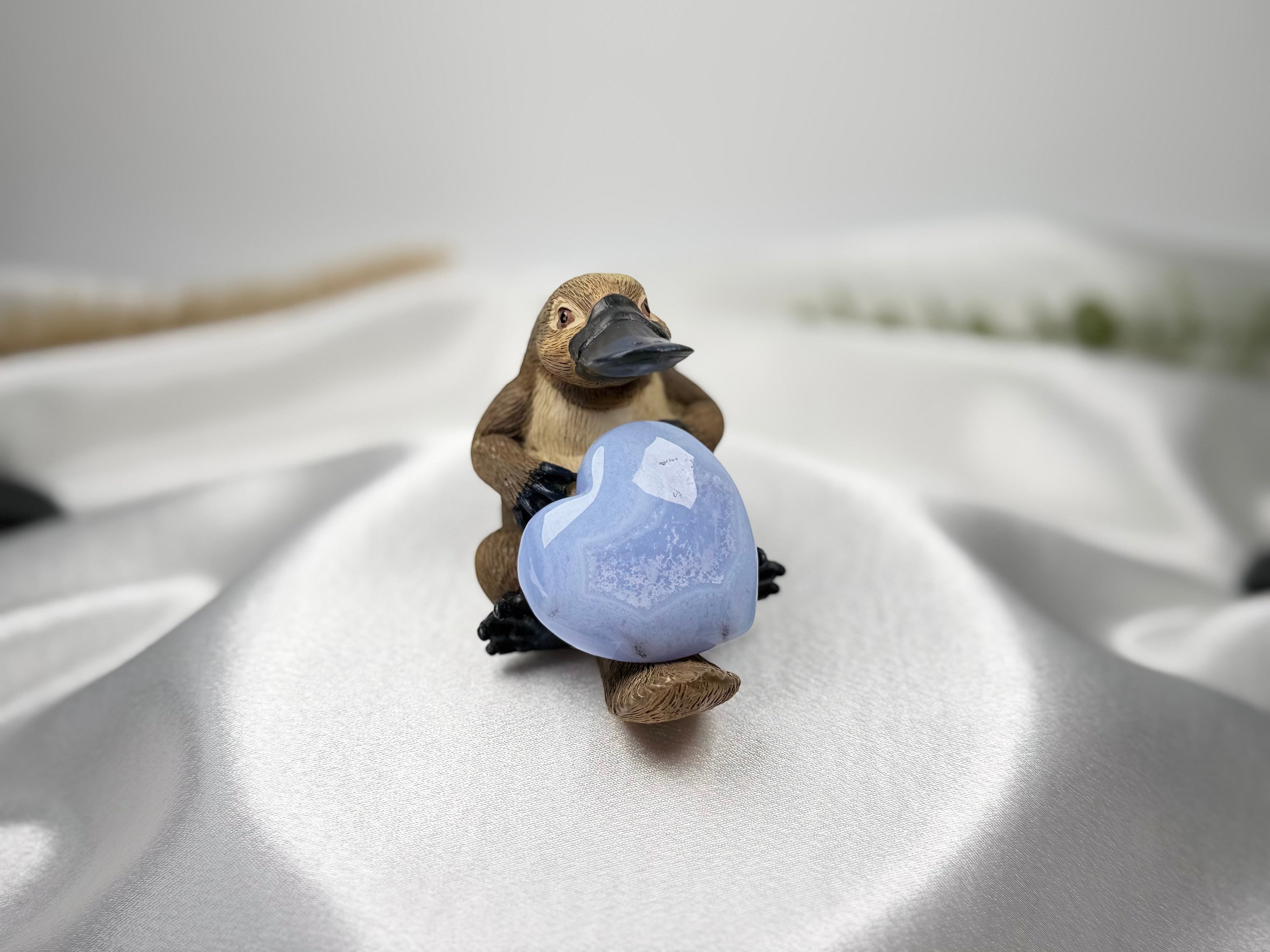 Treasured Energies Blue Lace Agate crystal heart held by a small platypus figurine on a soft white satin background.