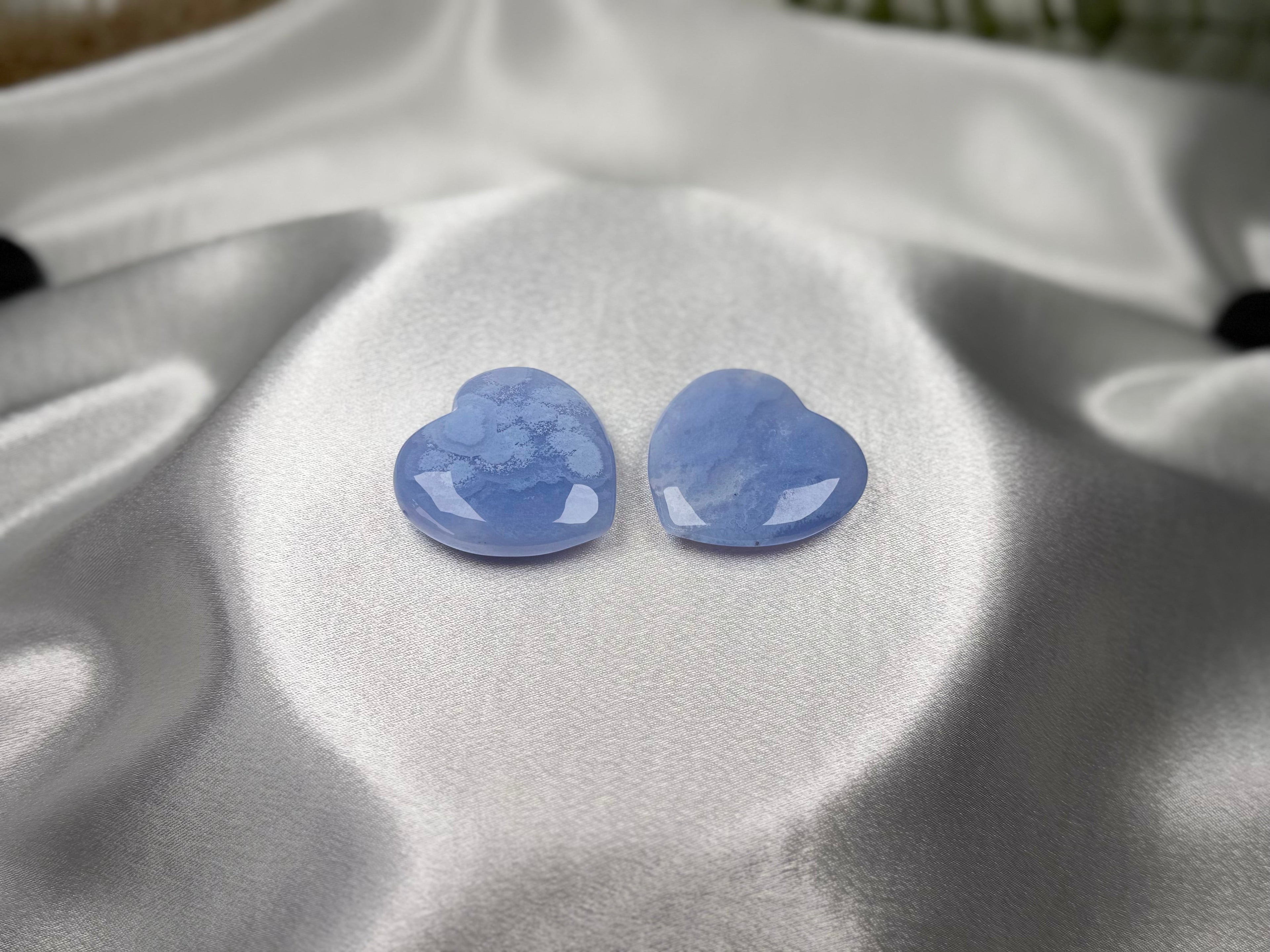 Two polished Blue Lace Agate crystal hearts from Treasured Energies displayed on smooth white satin fabric.