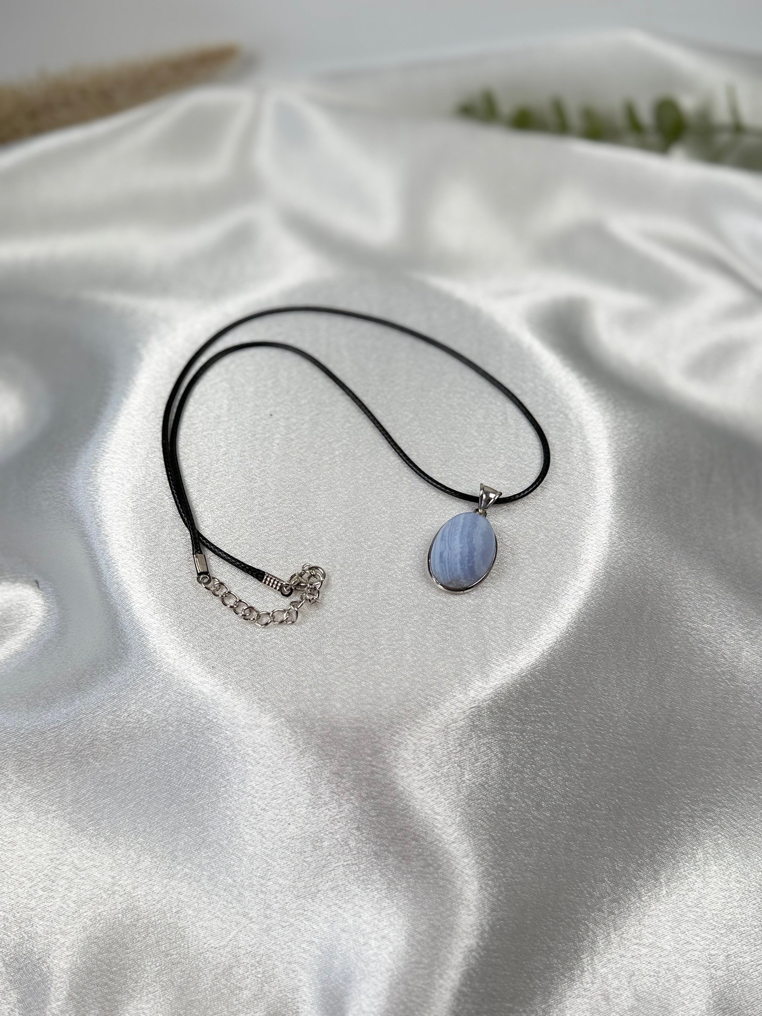 Treasured Energies blue lace agate pendant necklace with a matte finish on a black cord against white satin fabric.