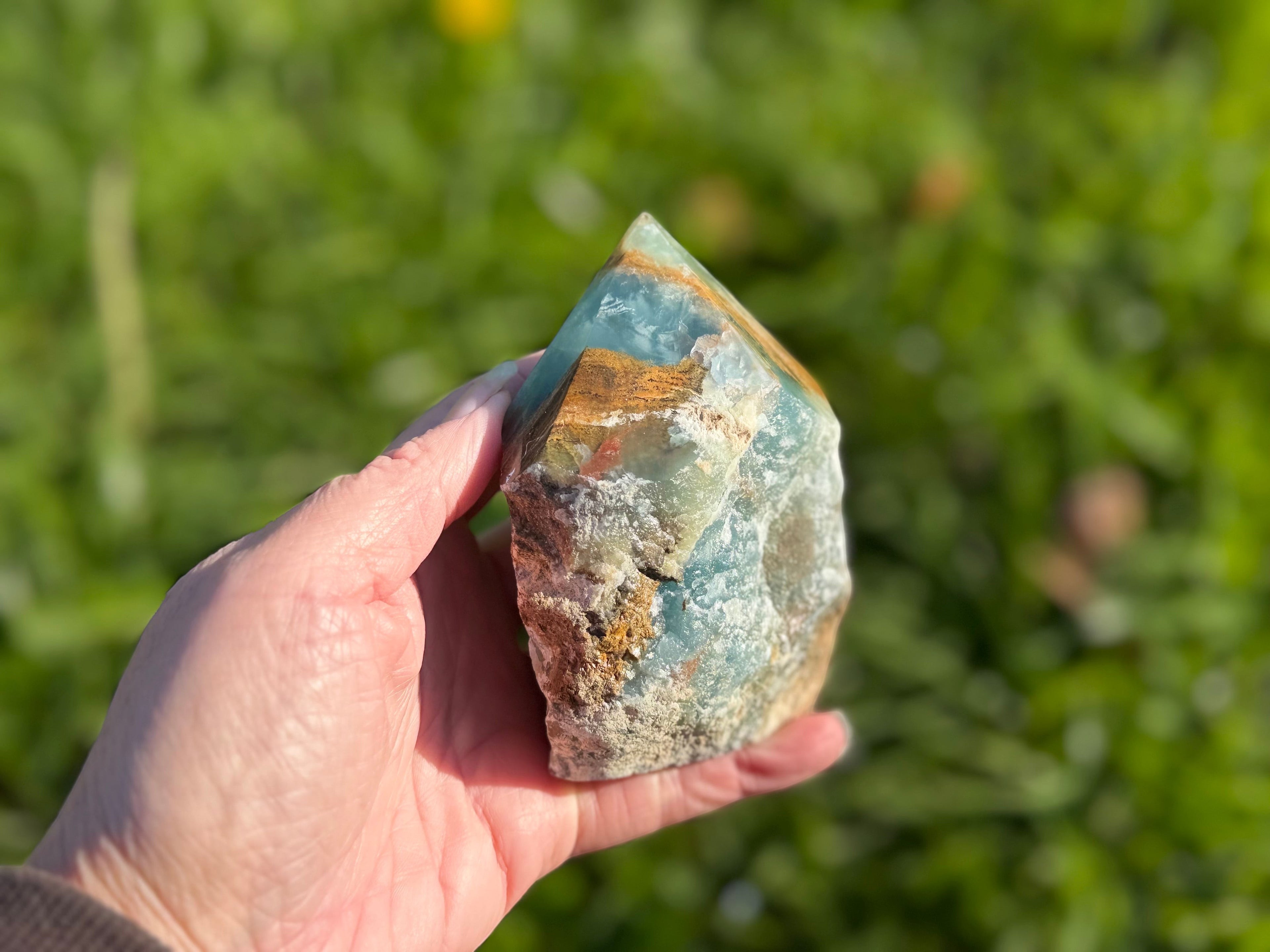 Treasured Energies blue onyx crystal tower with a raw, unpolished base held in a hand against a blurred green garden.
