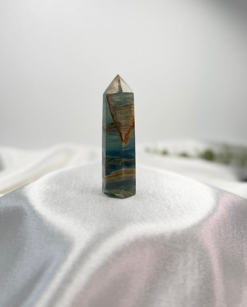 Treasured Energies polished Blue Onyx crystal tower with natural brown banding and a pointed tip on a white surface.