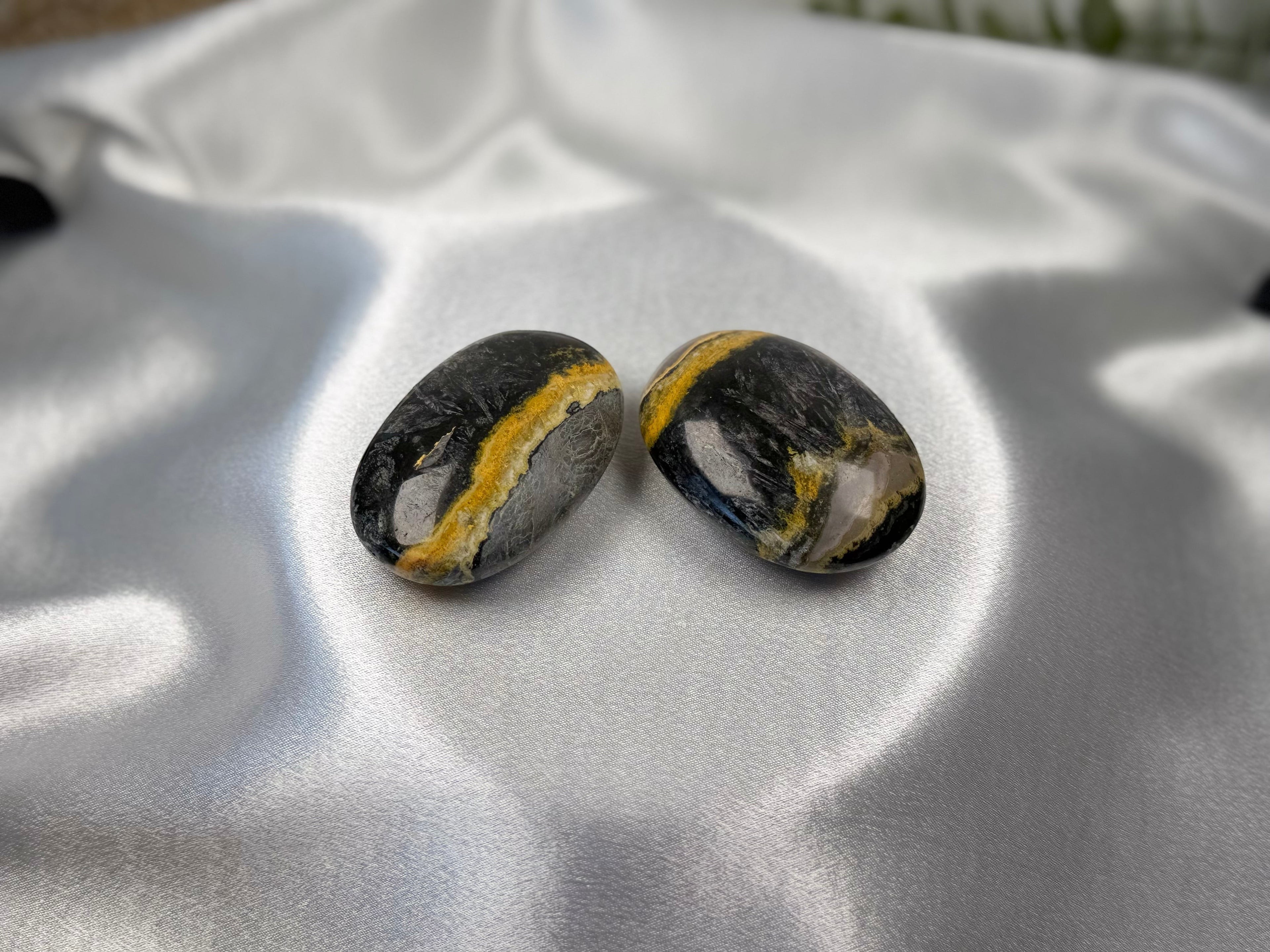 Two Treasured Energies polished Bumblebee Jasper palm stones with black, grey, and yellow bands on white satin fabric.