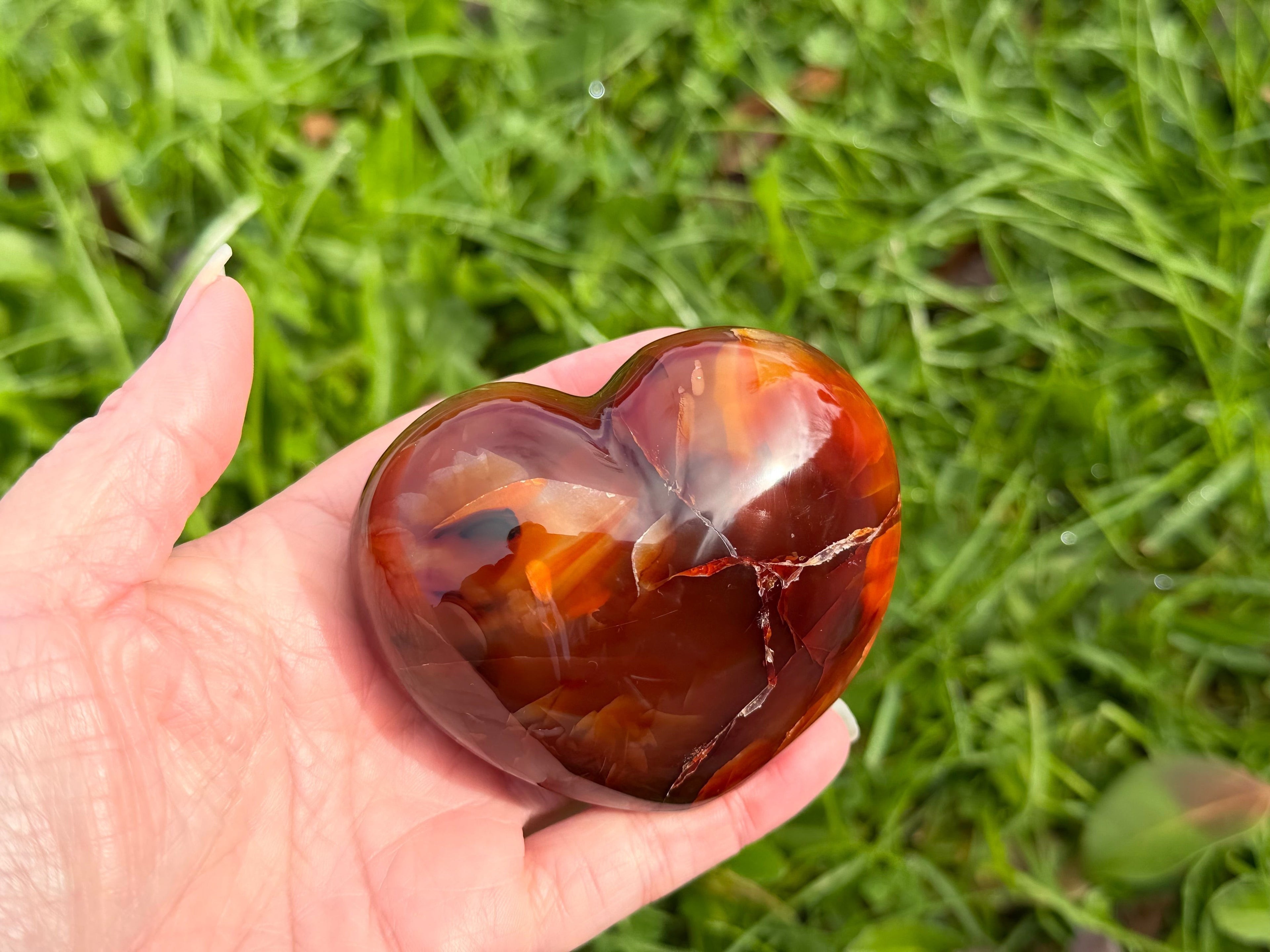 Treasured Energies polished carnelian crystal heart held in a hand against a green grass background