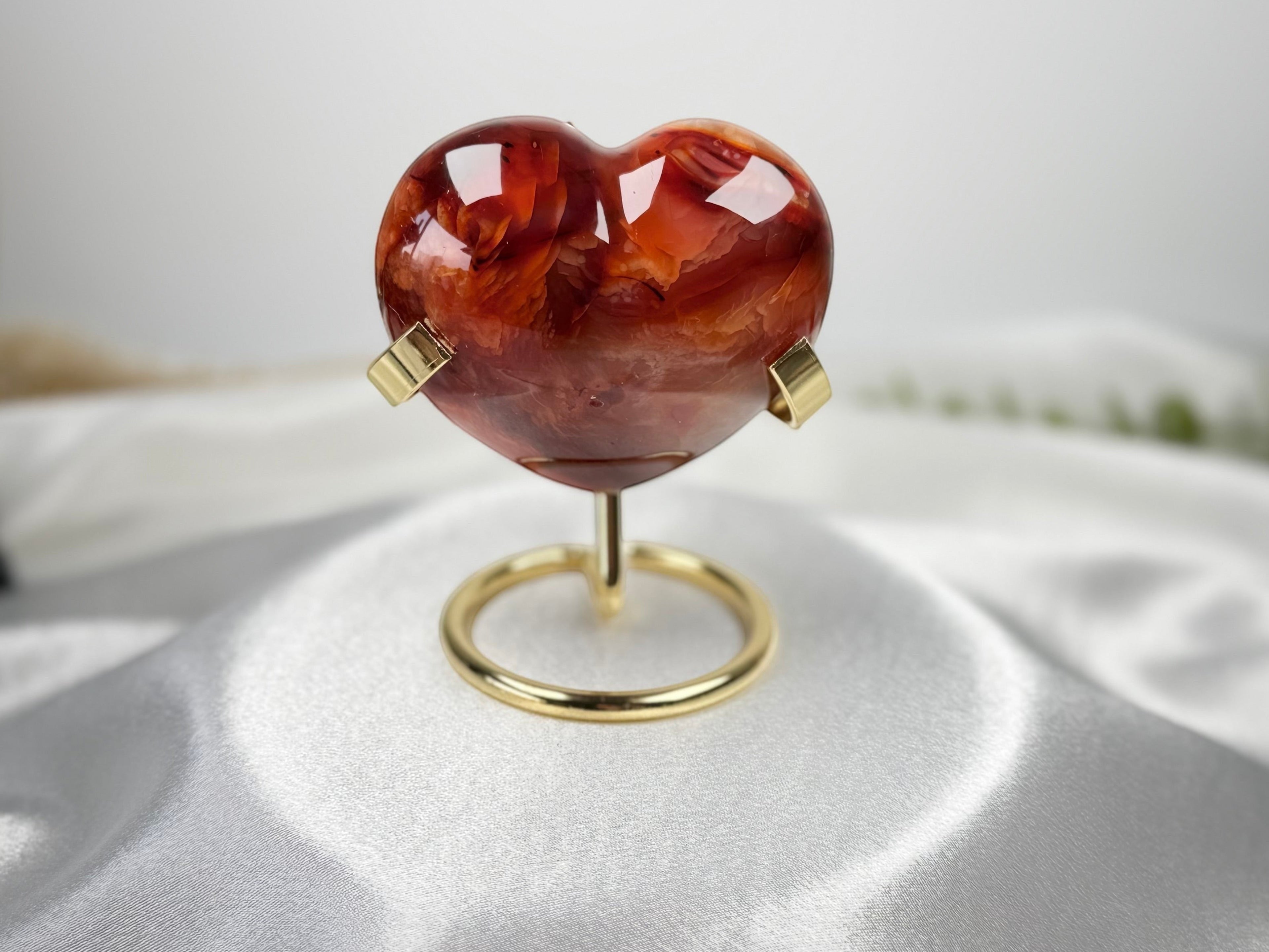 Treasured Energies polished carnelian crystal heart on a gold metal display stand against a white satin background.