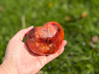 Treasured Energies large polished carnelian crystal heart held in a hand against a blurred green outdoor background