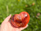 Treasured Energies large polished carnelian crystal heart held in a hand against a blurred green outdoor background