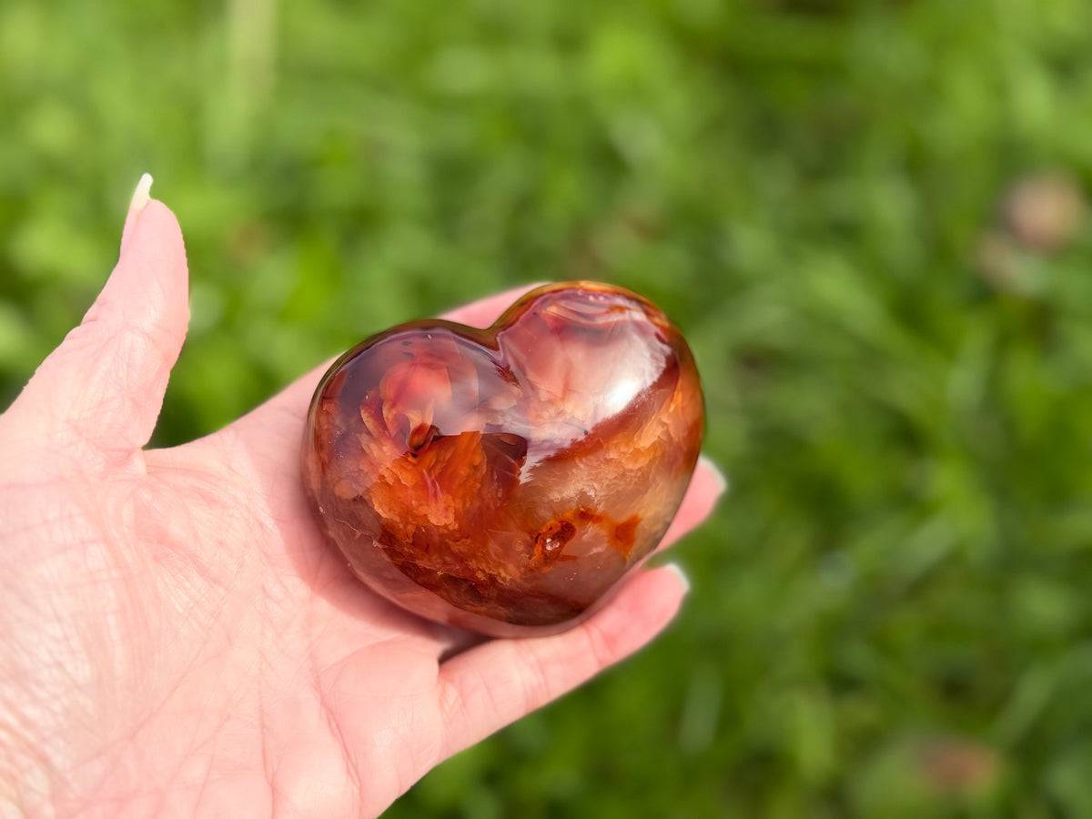 Treasured Energies polished carnelian crystal heart held in hand against a soft green garden background.
