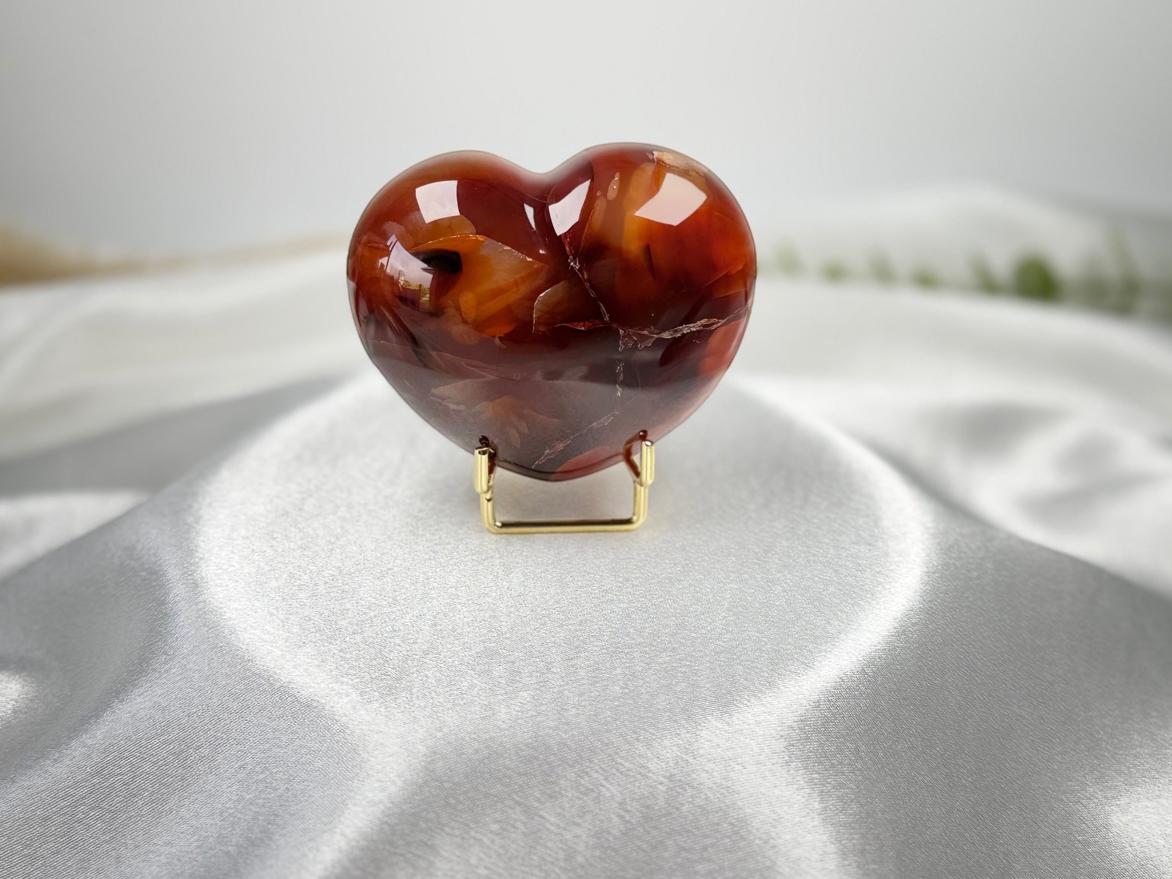 Treasured Energies polished carnelian crystal heart display piece on a gold stand against a white satin background.