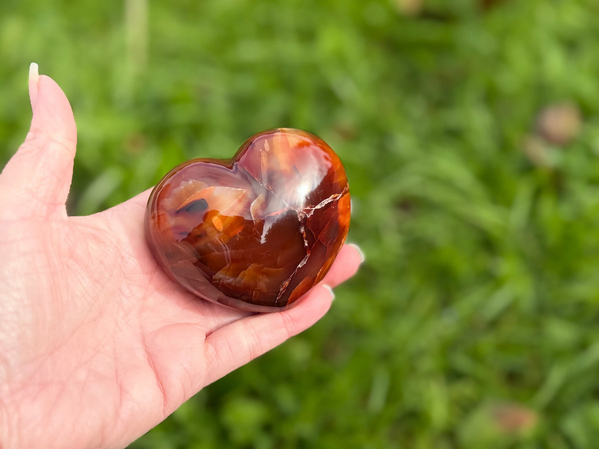 Treasured Energies polished carnelian crystal heart held in a hand against a blurred green grass background.