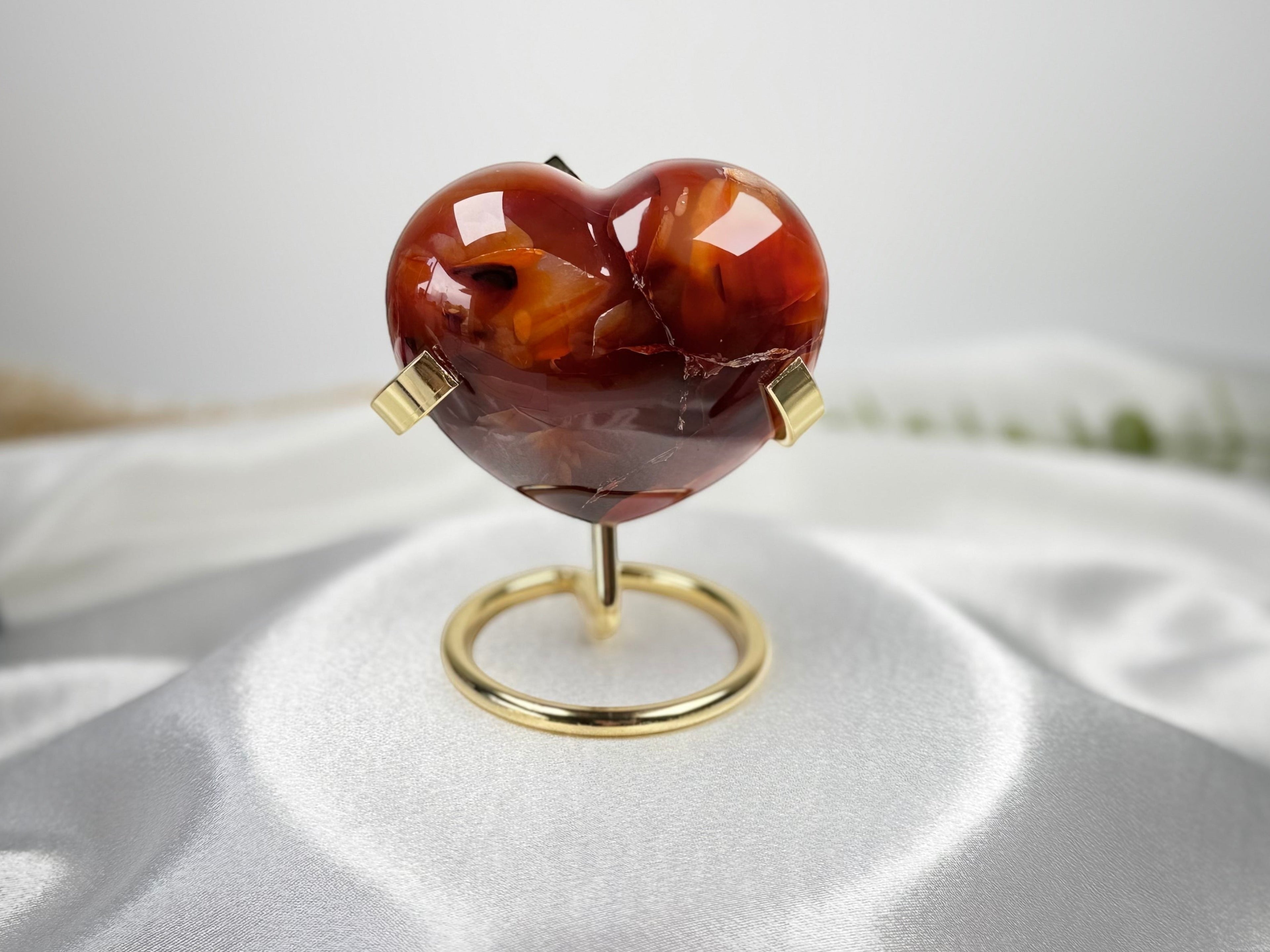 Treasured Energies polished carnelian crystal heart displayed on a gold metal stand against a white satin background.