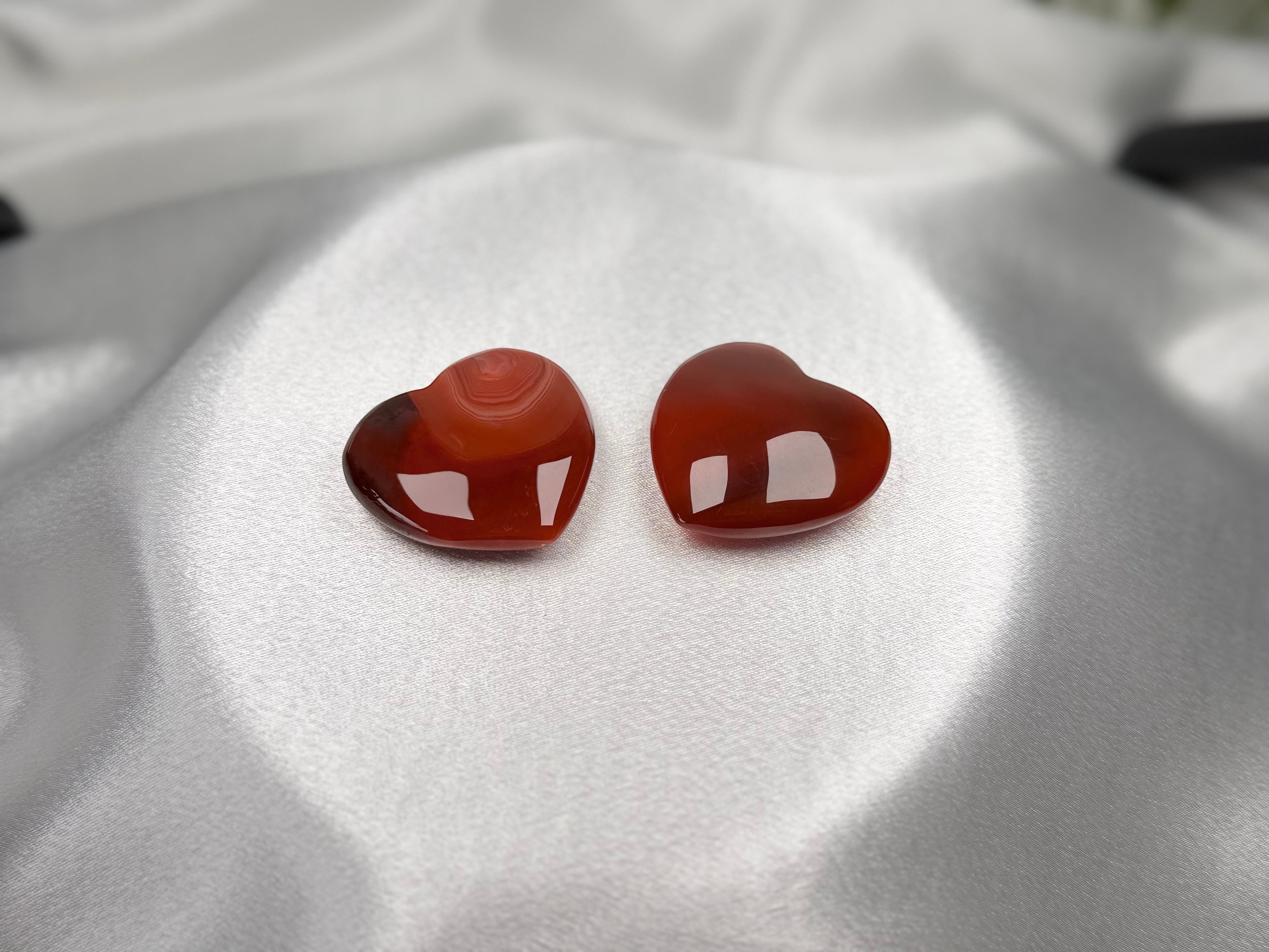 Two polished Treasured Energies carnelian heart crystals with deep orange and red banding on a white satin fabric.