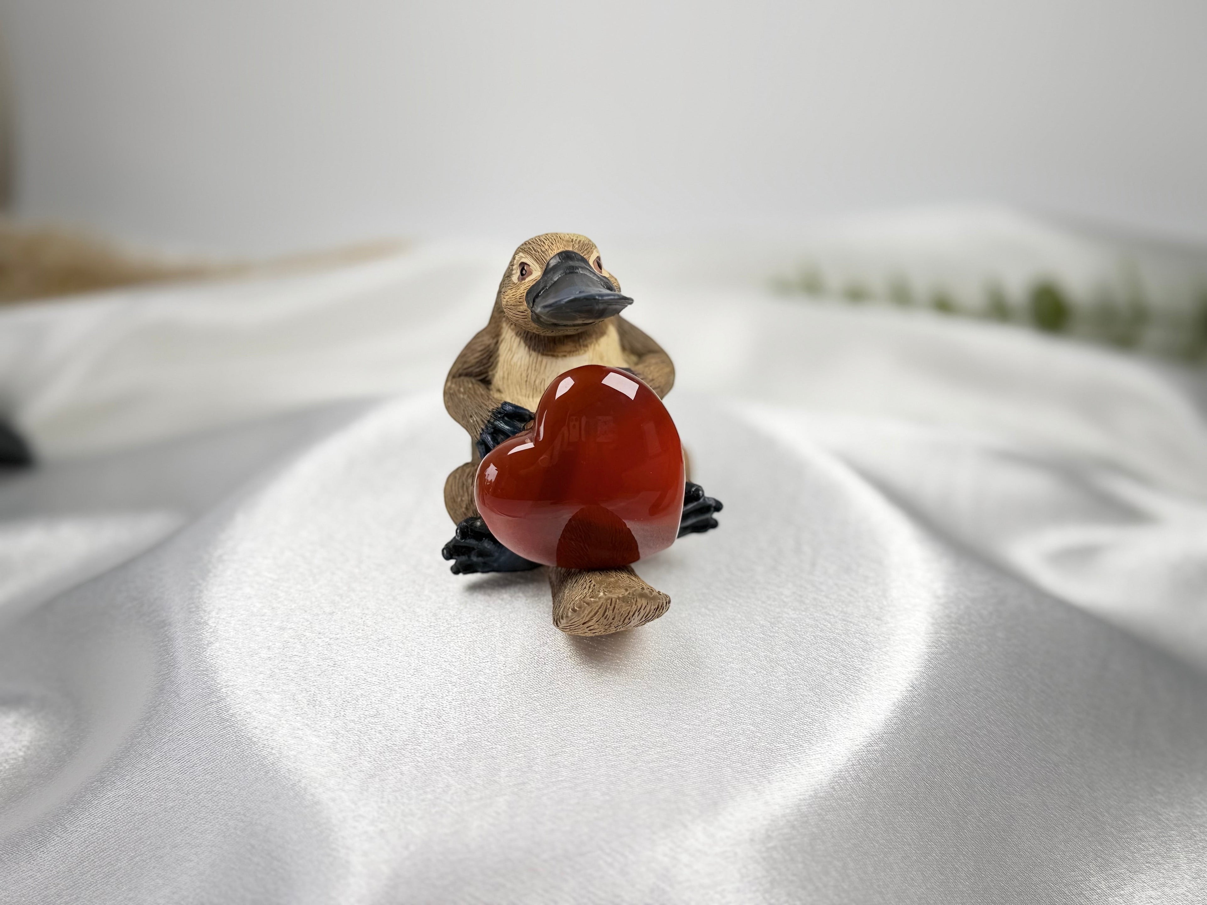 Treasured Energies polished carnelian heart held by a small platypus figurine on a white satin background.
