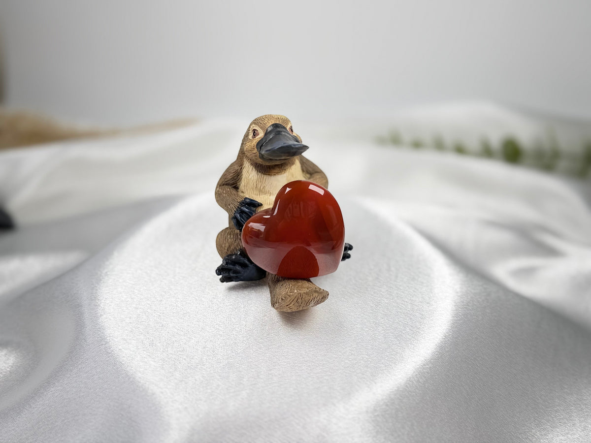 Treasured Energies carnelian heart held by a small, detailed platypus figurine on a white satin fabric background.