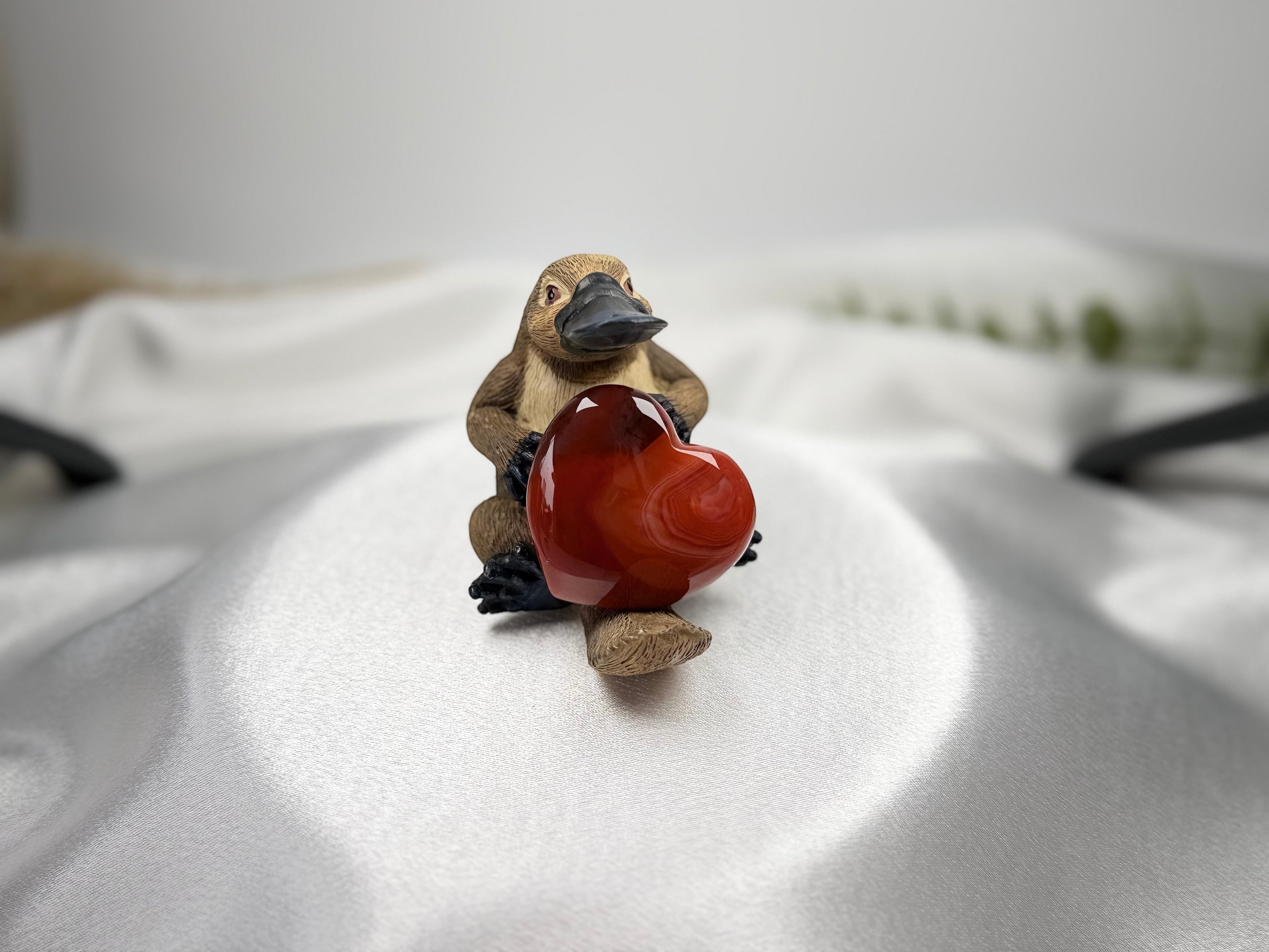 Treasured Energies carnelian heart held by a small carved wooden platypus figurine on a white satin background