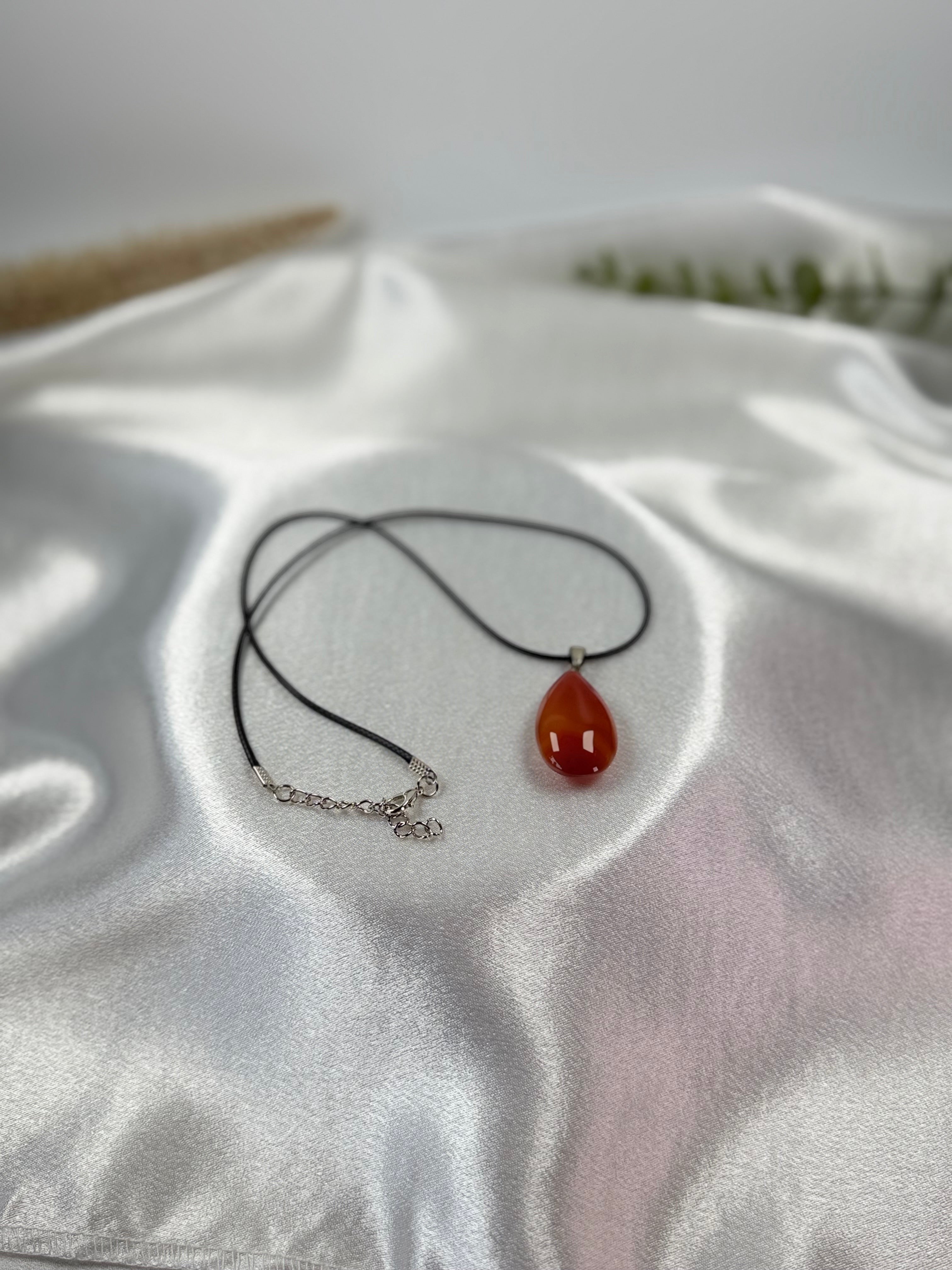 Treasured Energies carnelian teardrop gemstone pendant on a black cord necklace resting on white satin fabric.