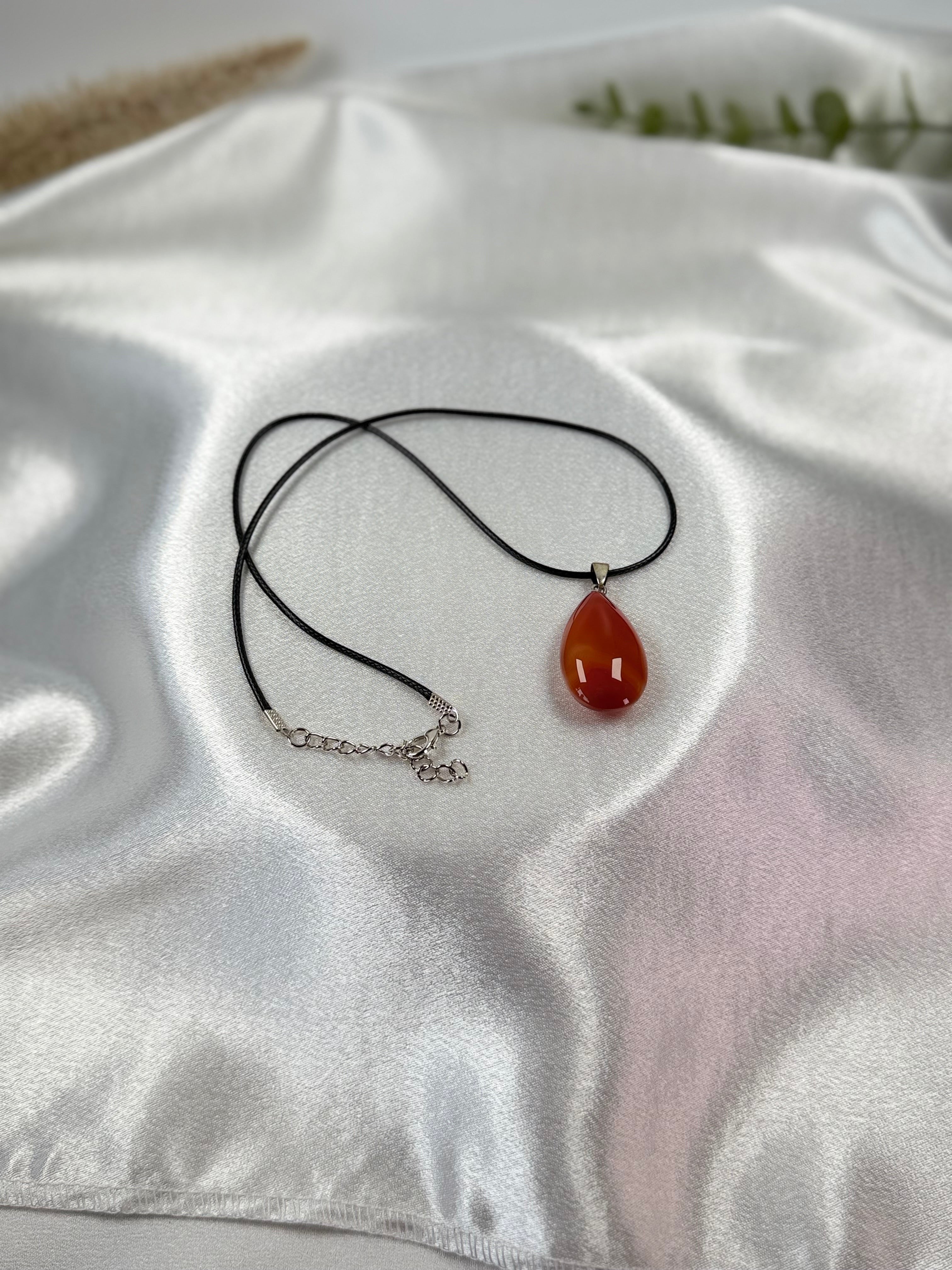 Treasured Energies carnelian teardrop gemstone pendant on a black cord necklace resting on white satin fabric.