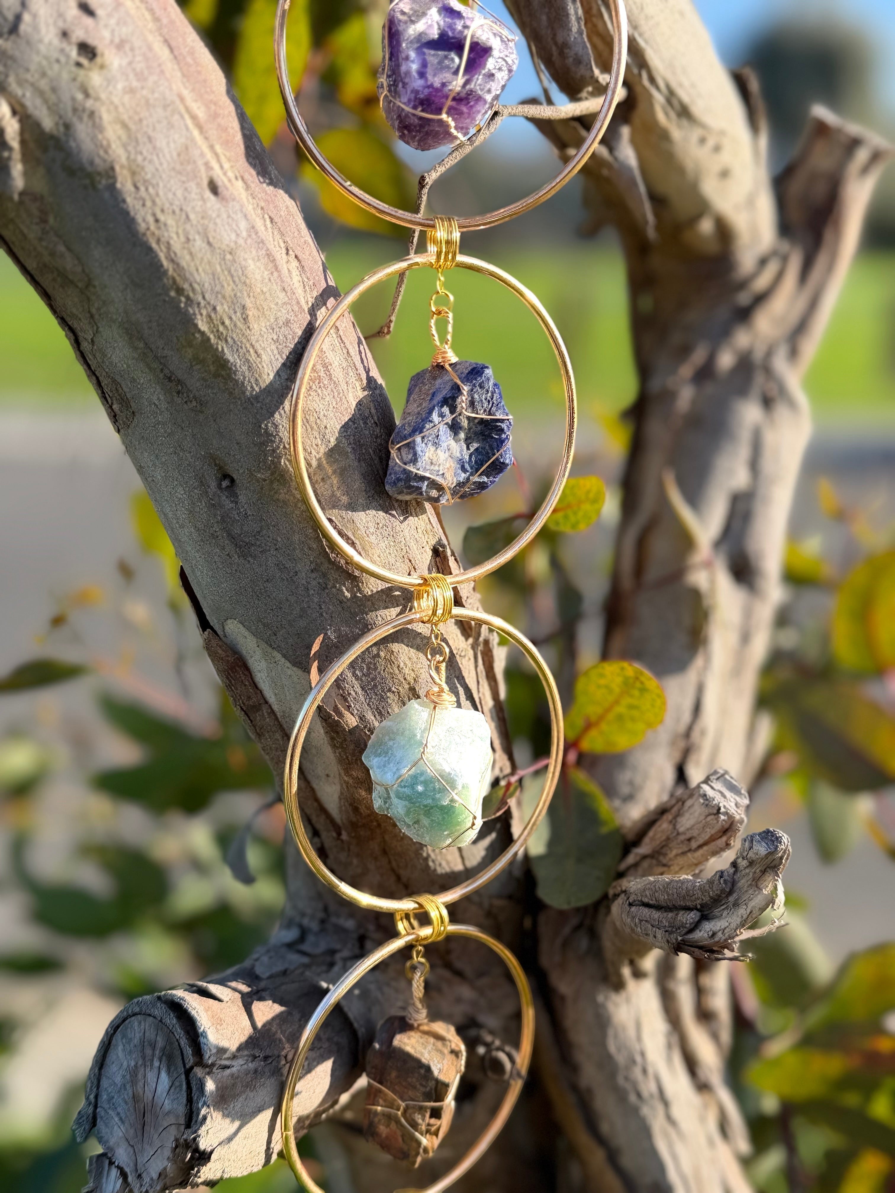 Treasured Energies 7 chakra crystal window hanging featuring raw gemstones in gold circles hanging on a tree branch.