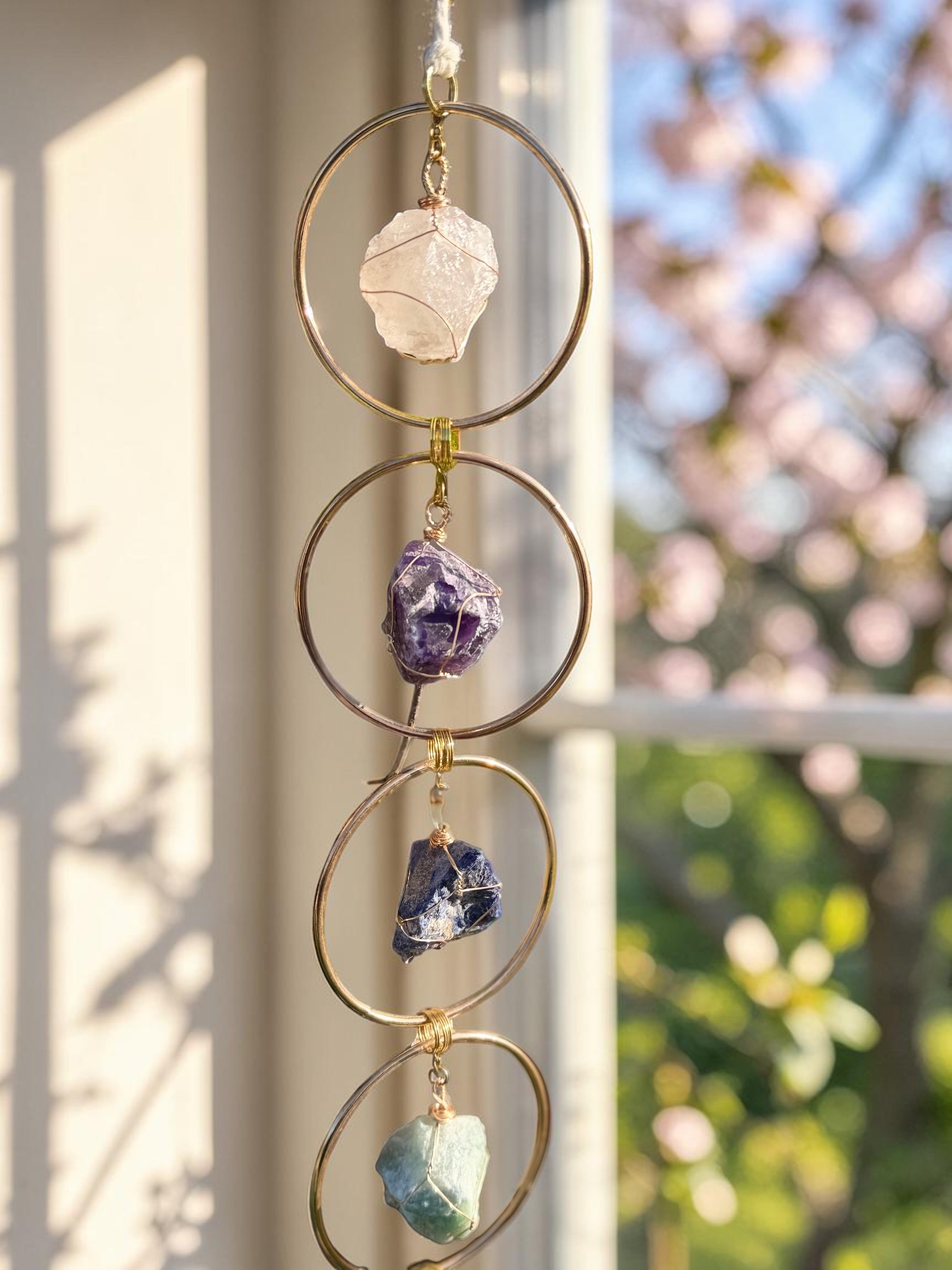 Treasured Energies 7 chakra crystal window hanging with raw gemstones in gold circles against a sunlit garden background.