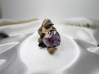 Treasured Energies Chevron Amethyst heart crystal held by a small platypus figurine on a white satin fabric background.