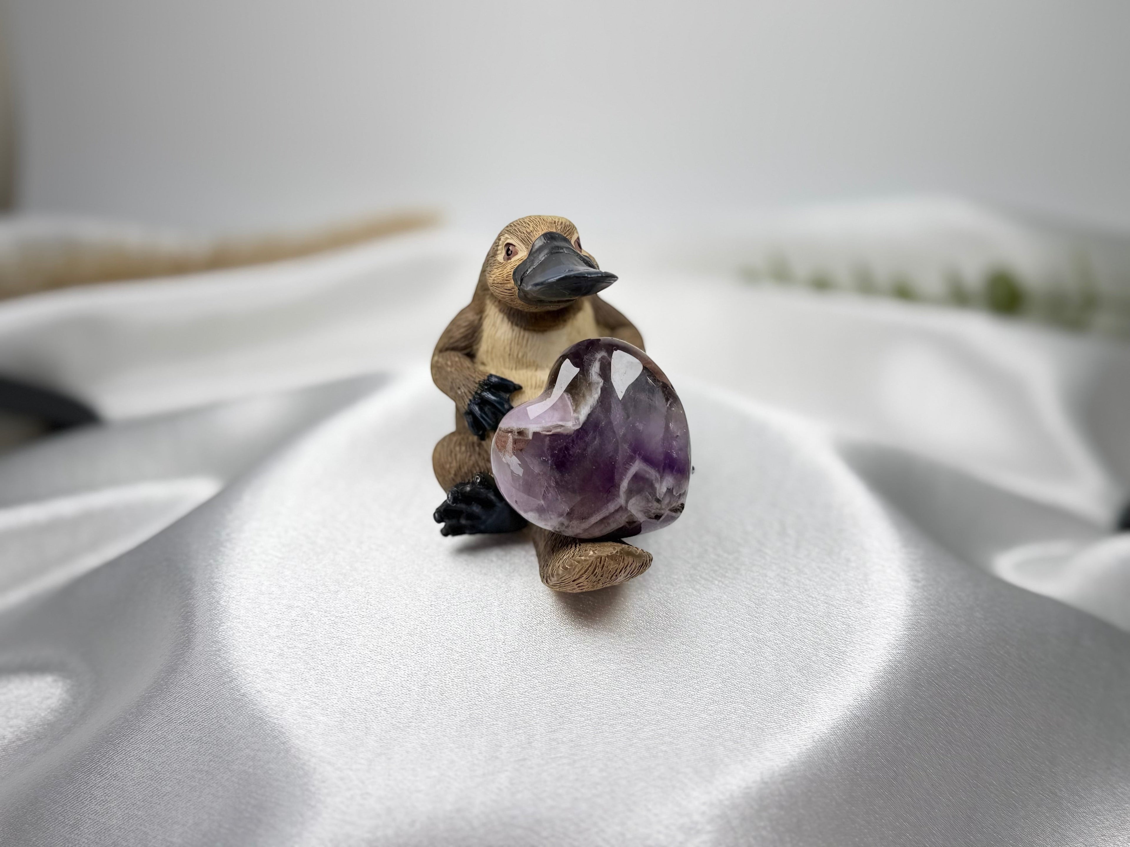 Treasured Energies Chevron Amethyst heart crystal held by a small platypus figurine on a white satin fabric background.