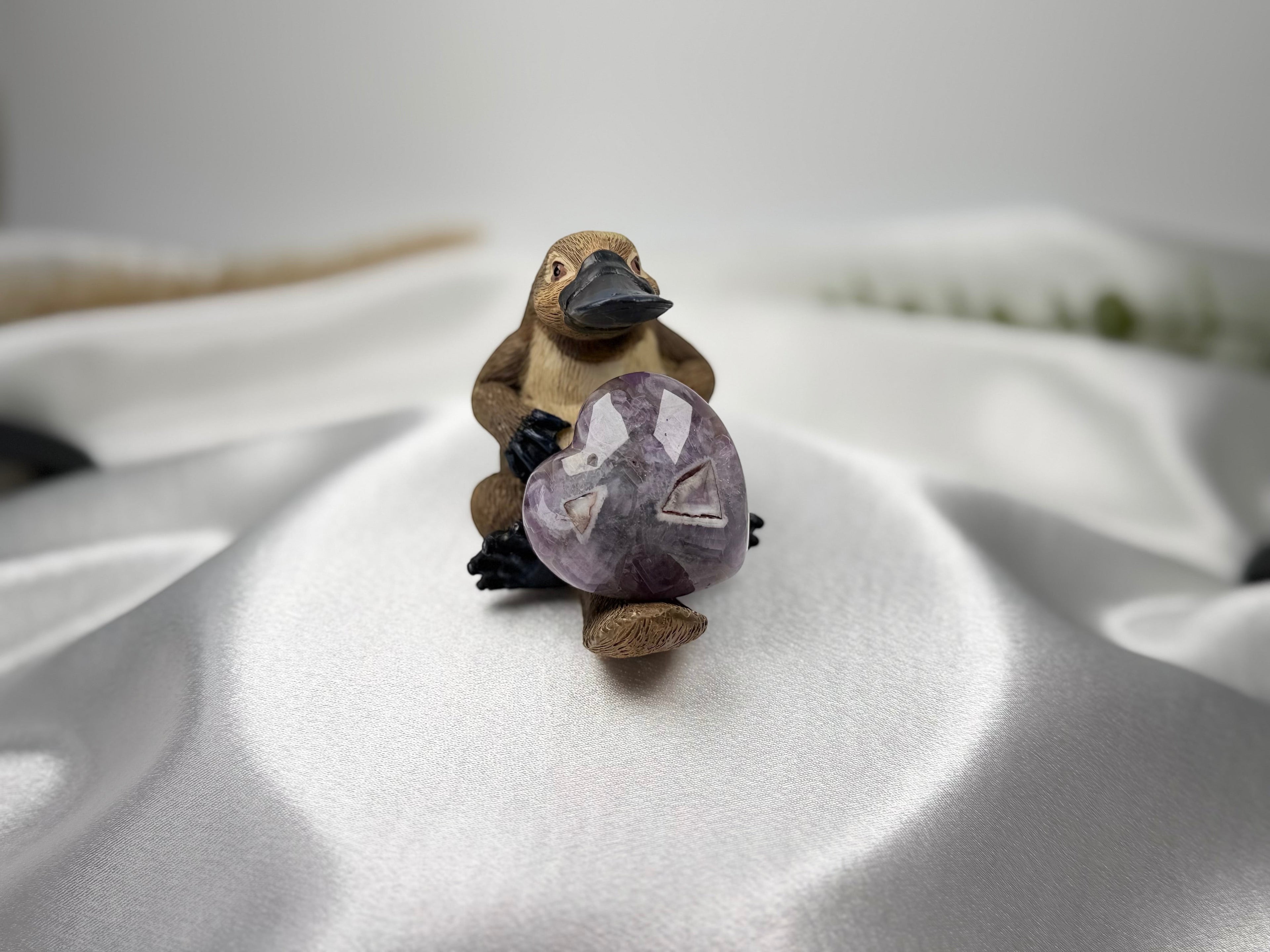Treasured Energies heart-shaped Chevron Amethyst crystal held by a small platypus figurine on white satin fabric.