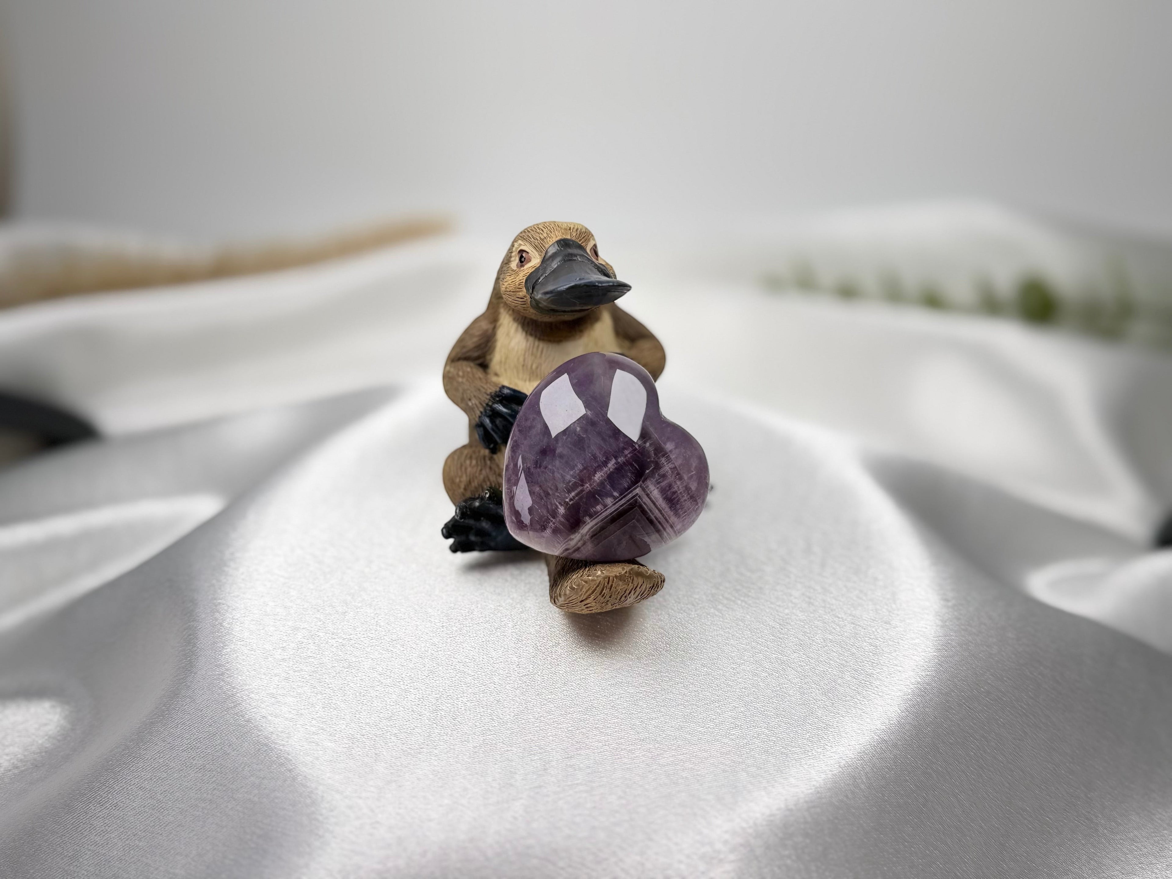 Treasured Energies Chevron Amethyst heart crystal held by a small platypus figurine on a white satin background.
