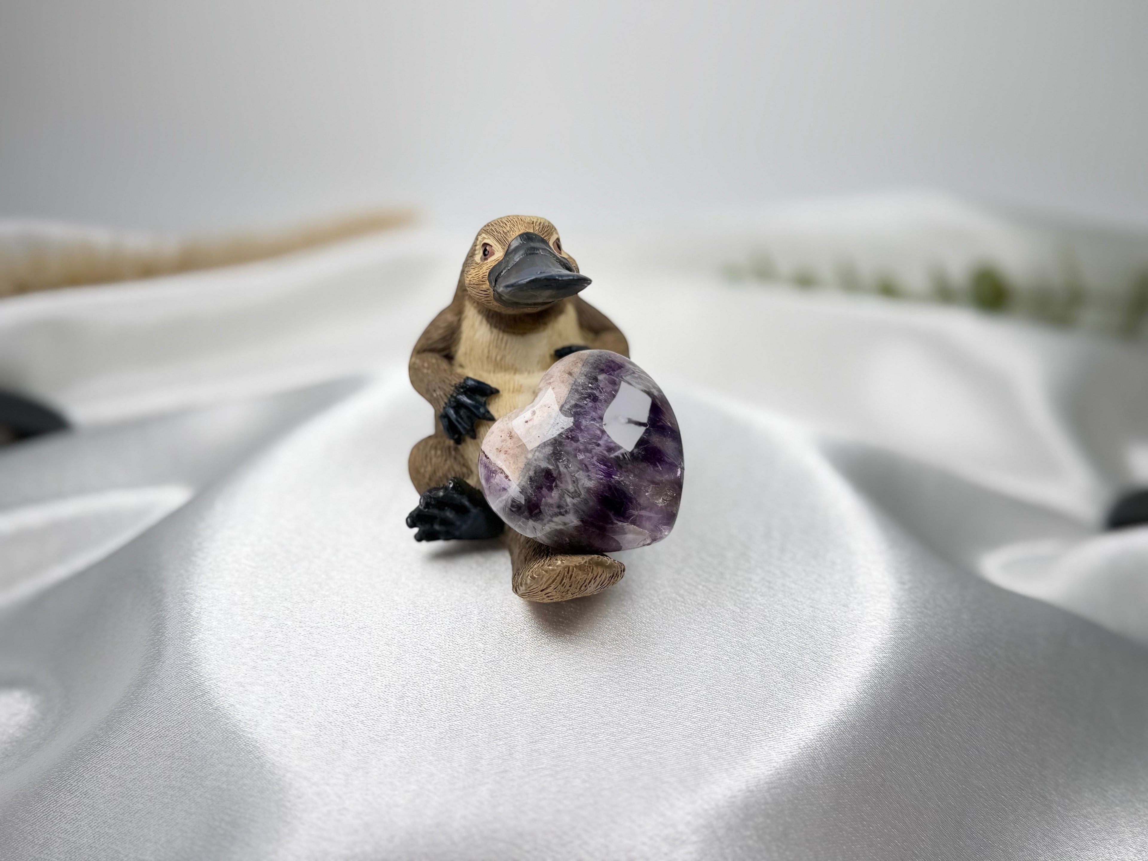 Treasured Energies heart-shaped Chevron Amethyst crystal held by a small platypus figurine on white satin fabric.