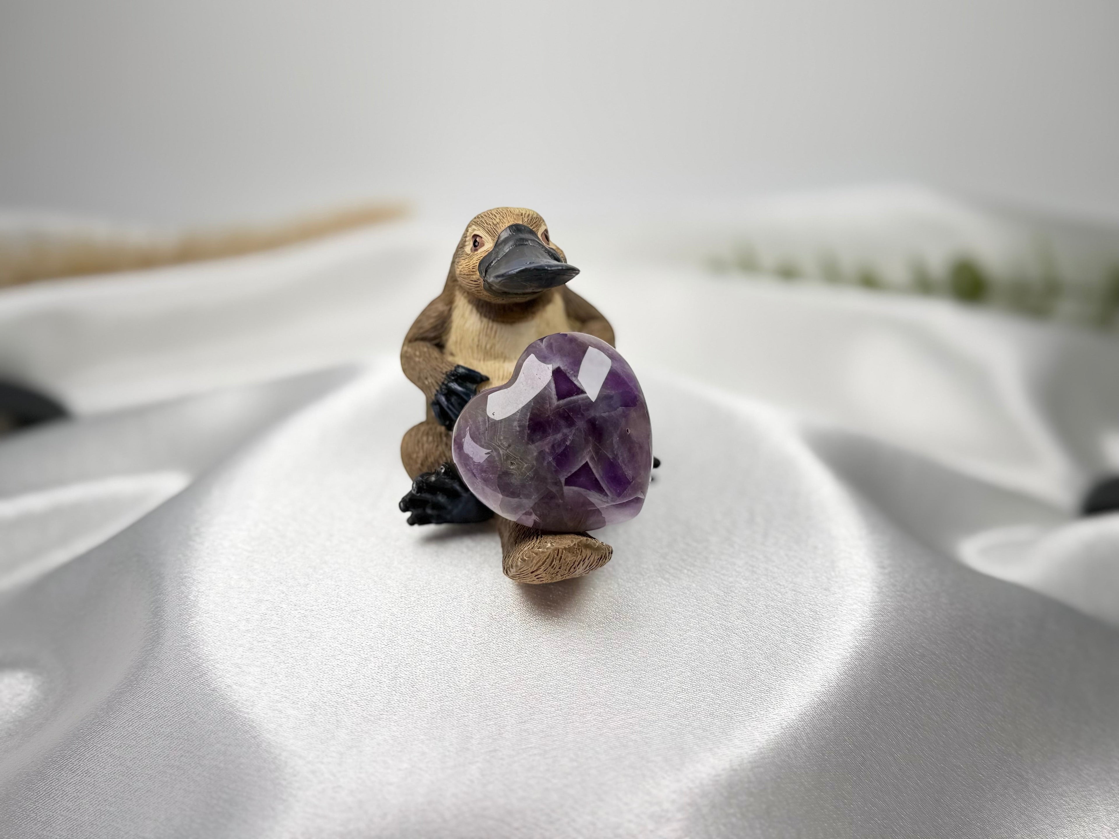 Treasured Energies heart-shaped Chevron Amethyst crystal held by a small platypus figurine on a white satin fabric