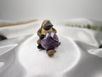 Treasured Energies heart-shaped Chevron Amethyst crystal held by a small platypus figurine on white satin fabric.