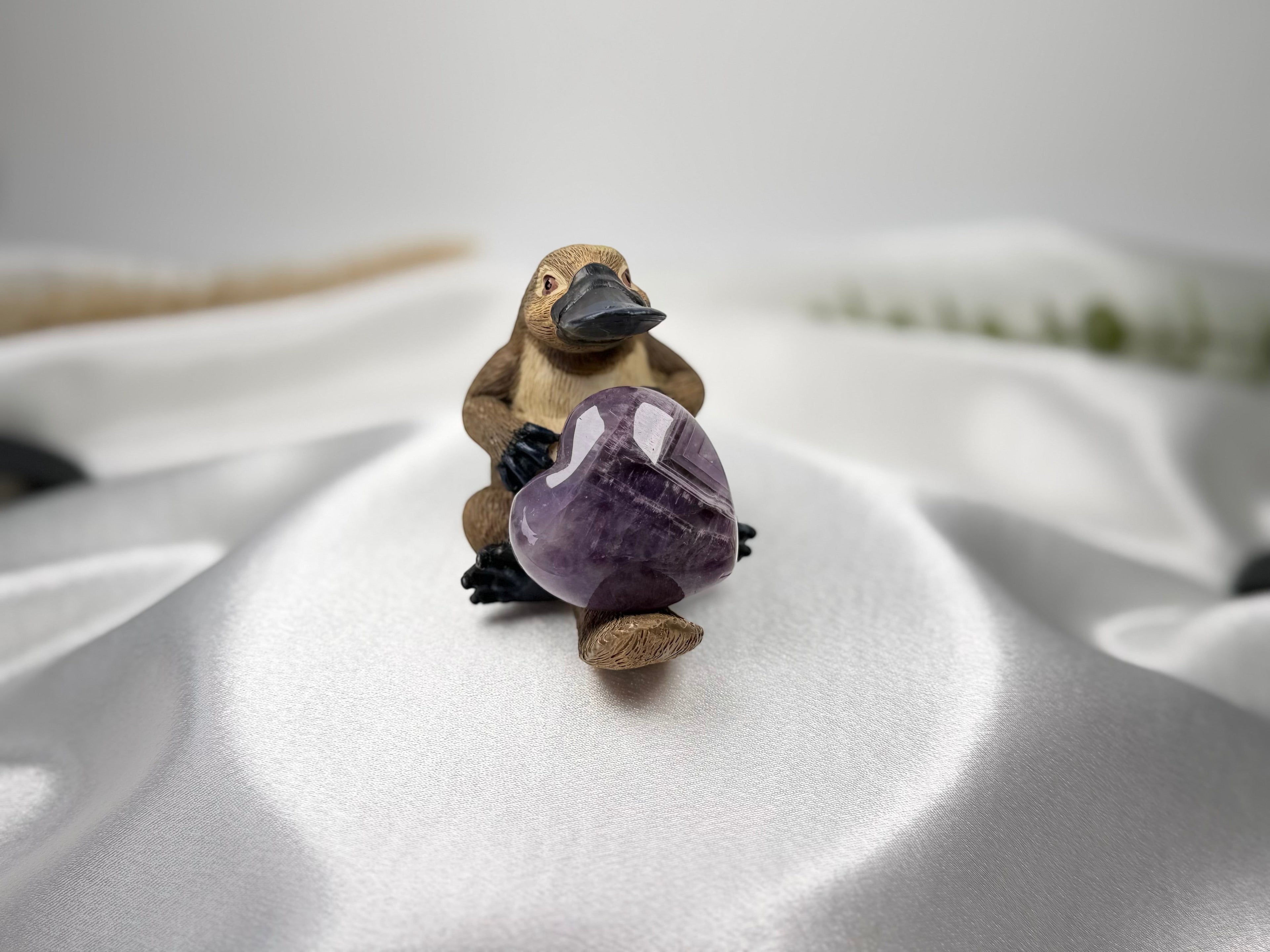 Treasured Energies heart-shaped Chevron Amethyst crystal held by a small platypus figurine on white satin fabric.