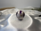 Treasured Energies polished Chevron Amethyst crystal sphere with purple and white V-shaped bands on white satin fabric.