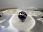 Treasured Energies polished Chevron Amethyst crystal sphere with deep purple and white V-shaped bands on satin fabric.