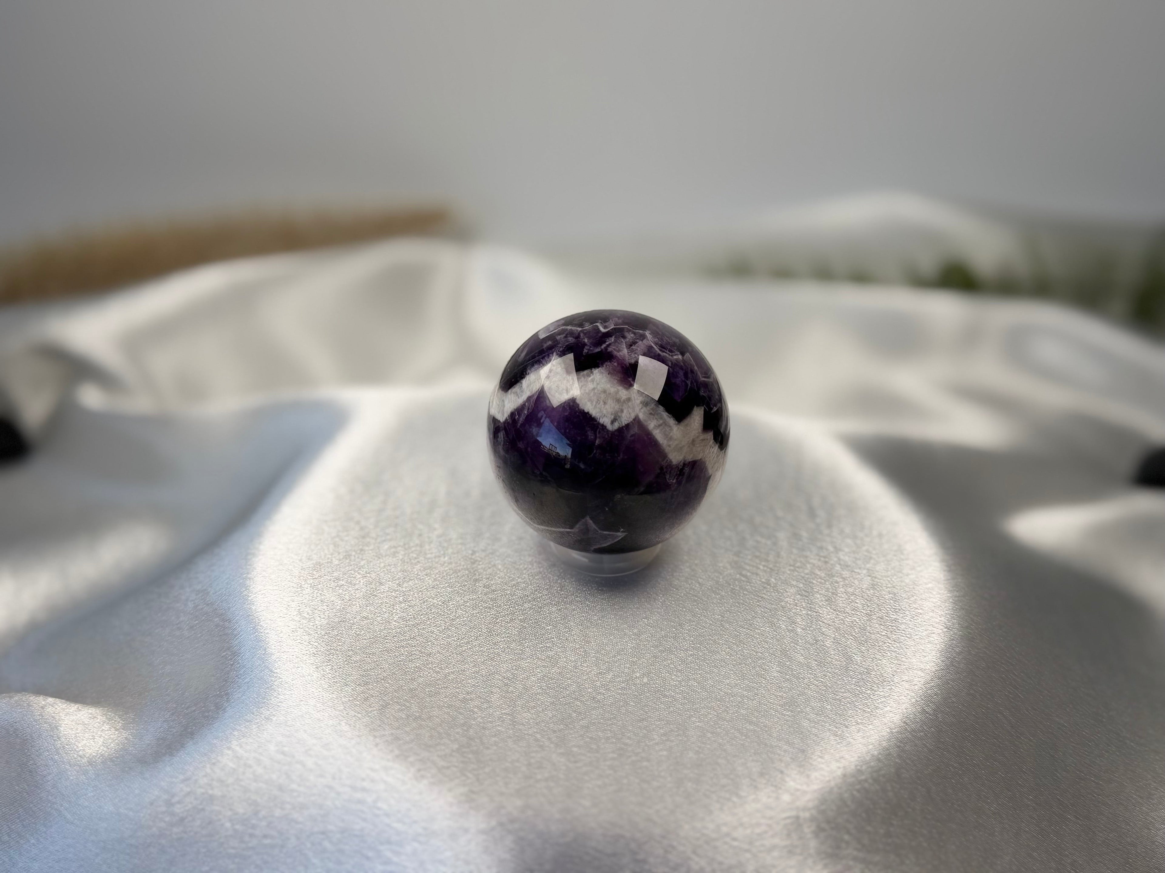 Treasured Energies polished Chevron Amethyst crystal sphere with deep purple and white V-shaped bands on satin fabric.