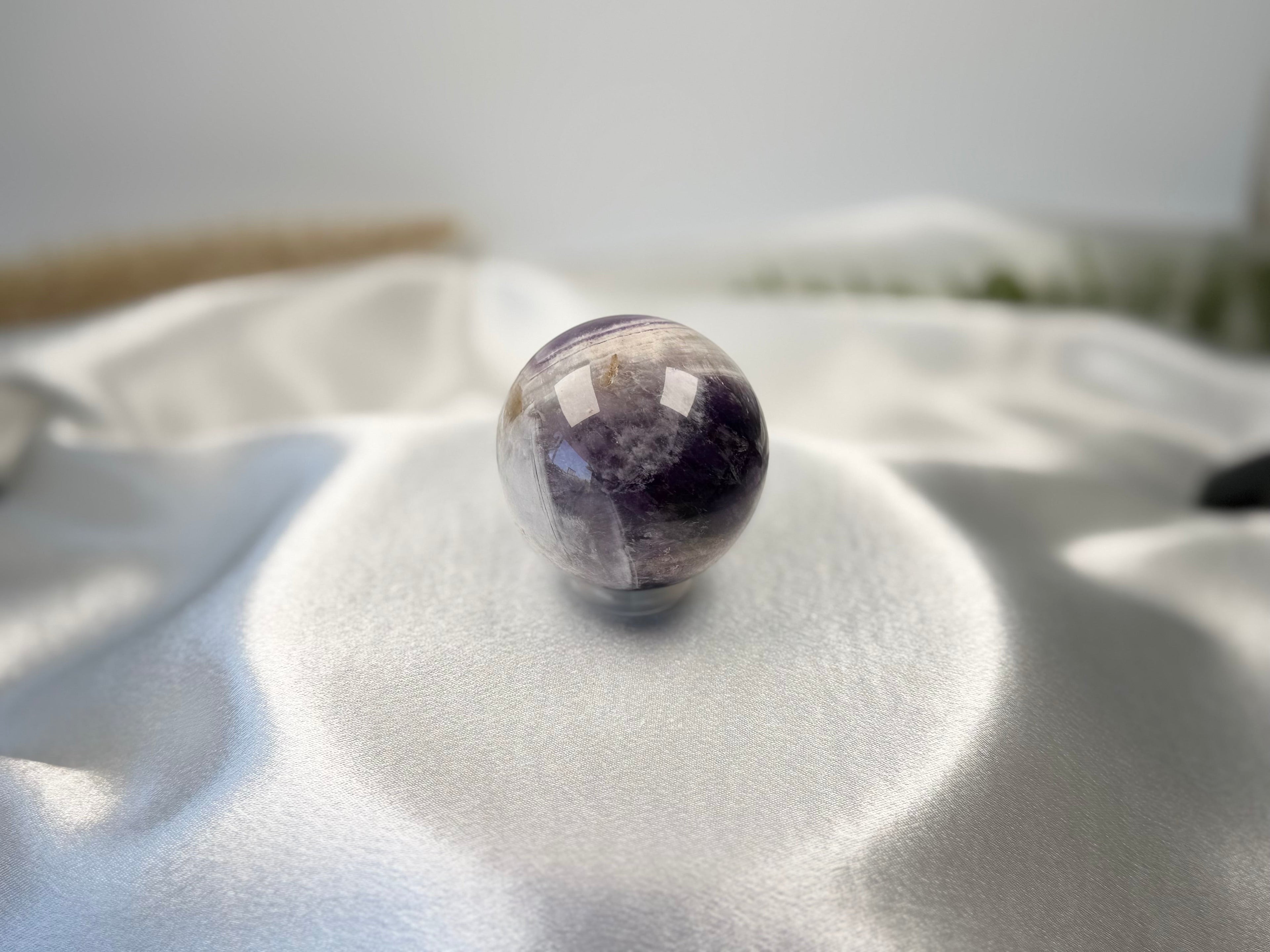 Treasured Energies polished Chevron Amethyst crystal sphere with purple and white V-shaped bands on white silk fabric.